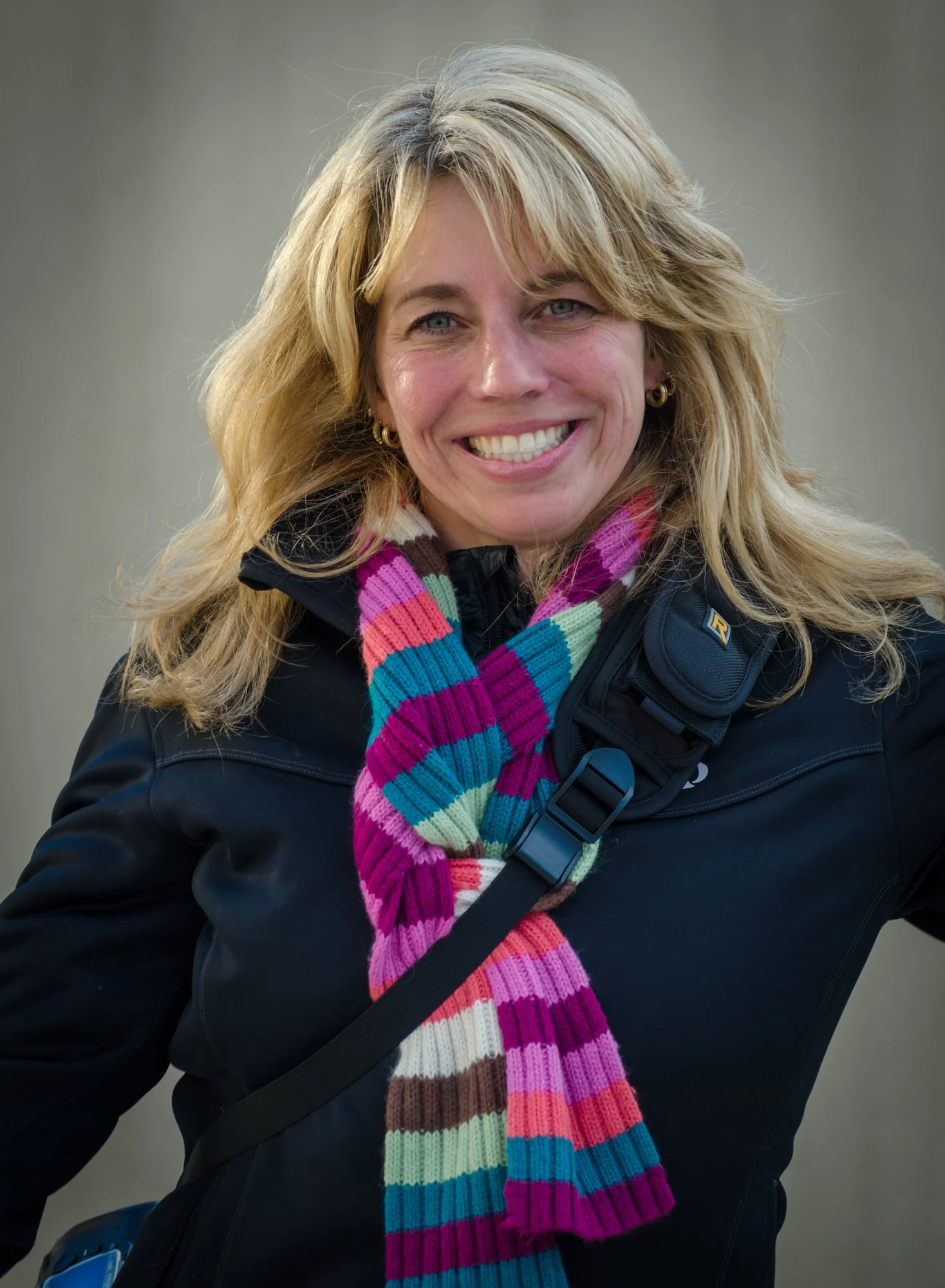 A woman with blonde hair, wearing a striped colorful scarf, a black jacket, and gold earrings, smiling outdoors with a blurred background.