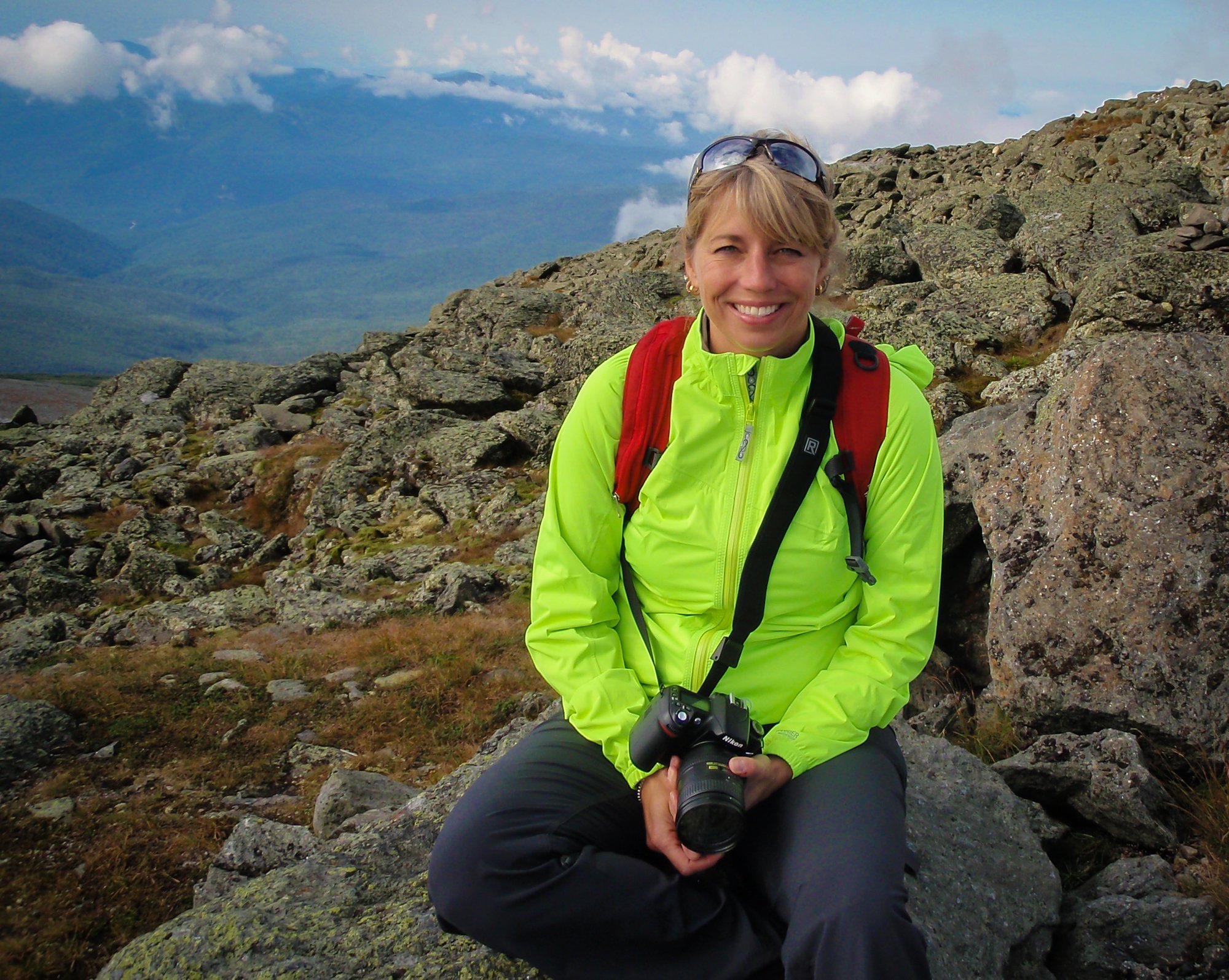Smiling woman in bright yellow outdoor jacket and sunglasses on her head sitting among rocks on a mountain trail, holding a Nikon camera, with a mountainous landscape and cloudy sky in the background.