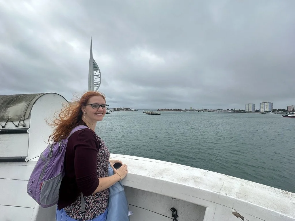 Smiling young woman with red hair and glasses standing on a boat, ocean in the background with a city skyline and iconic sail-shaped building under cloudy skies.