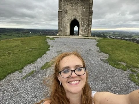 A woman with glasses and red hair taking a selfie in front of an old stone tower on a grassy hill, with a cloudy sky in the background.