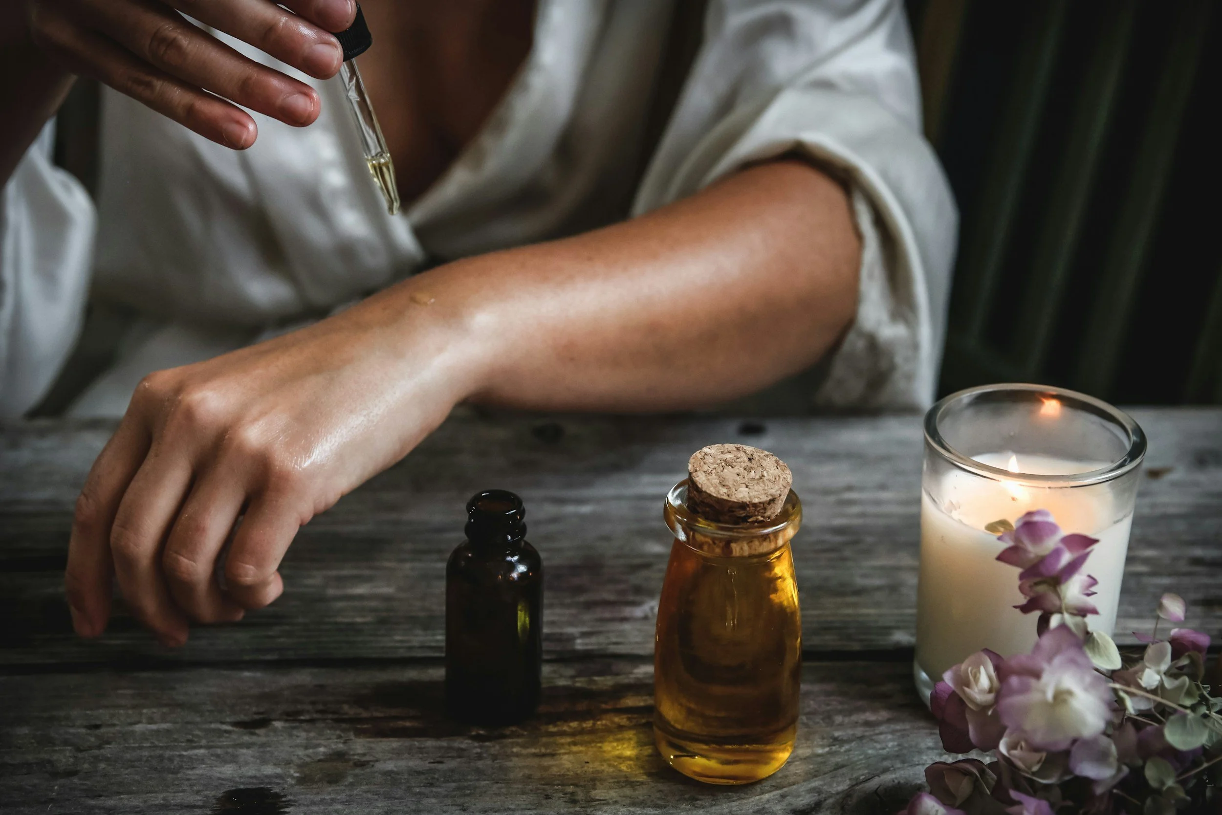 A person is using a dropper to transfer liquid into a small brown glass bottle. There are other bottles on the dark wooden table, a lit candle in a glass holder, and a bunch of pink and white flowers nearby.