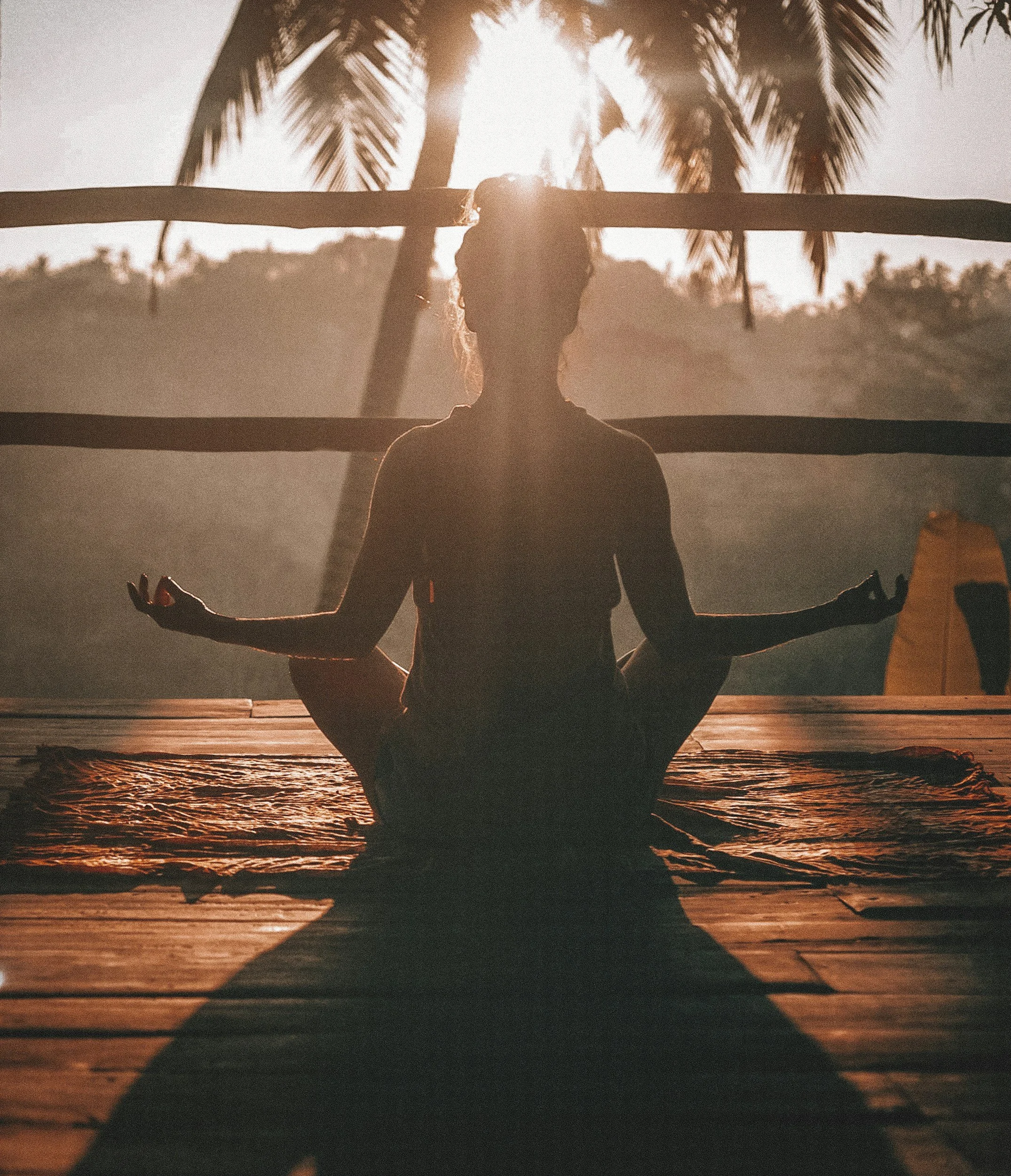 Silhouette of a person practicing yoga or meditation on a wooden deck during sunset at the beach, with palm trees in the background.