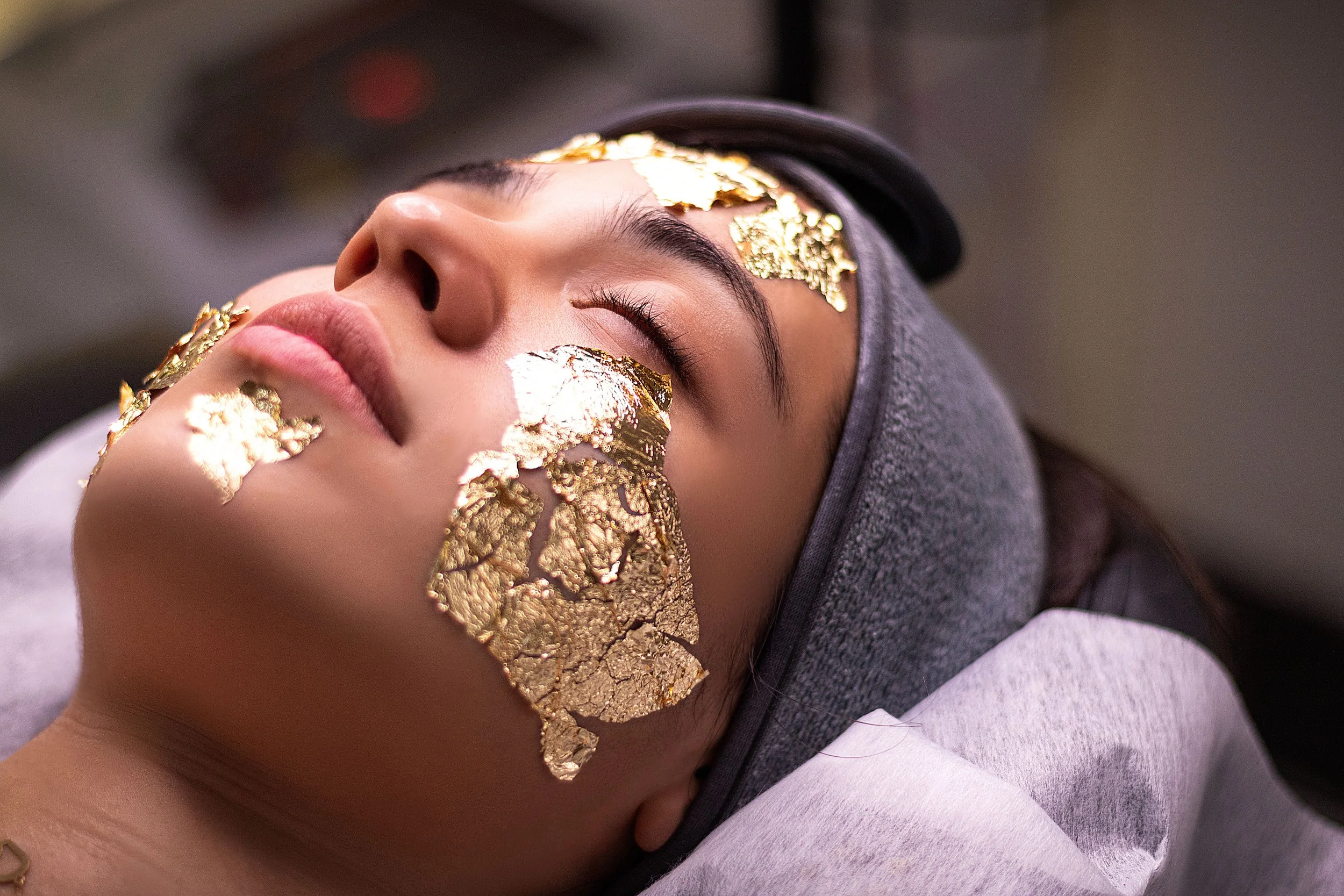 A woman lying down with her eyes closed, covered in gold leaf on her face, eyebrows, and cheek, in a skincare or spa treatment setting.