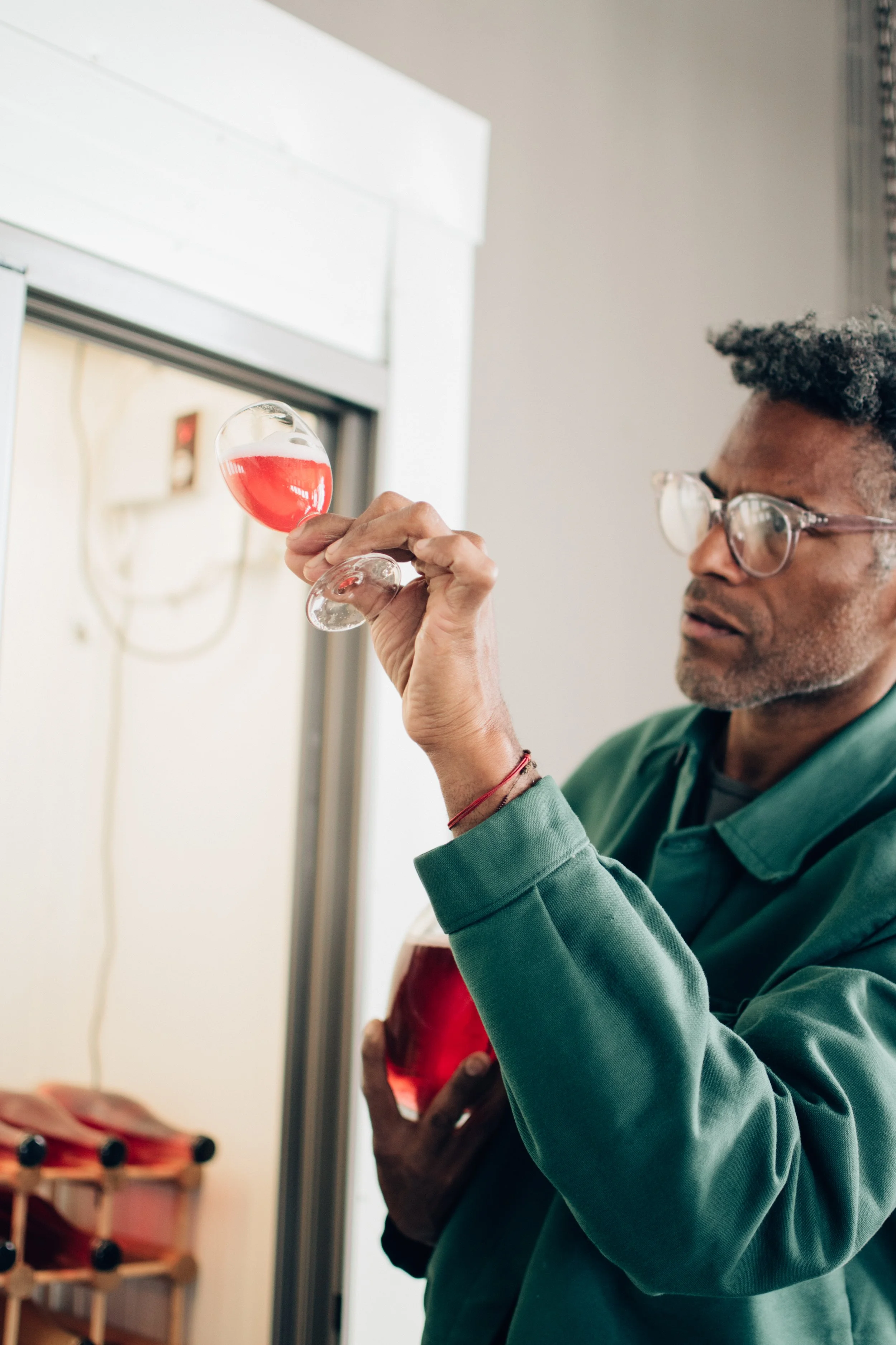 A person with glasses holding a wine glass filled with red scobybaby kombucha, standing indoors near a wine cooler or beverage refrigerator.