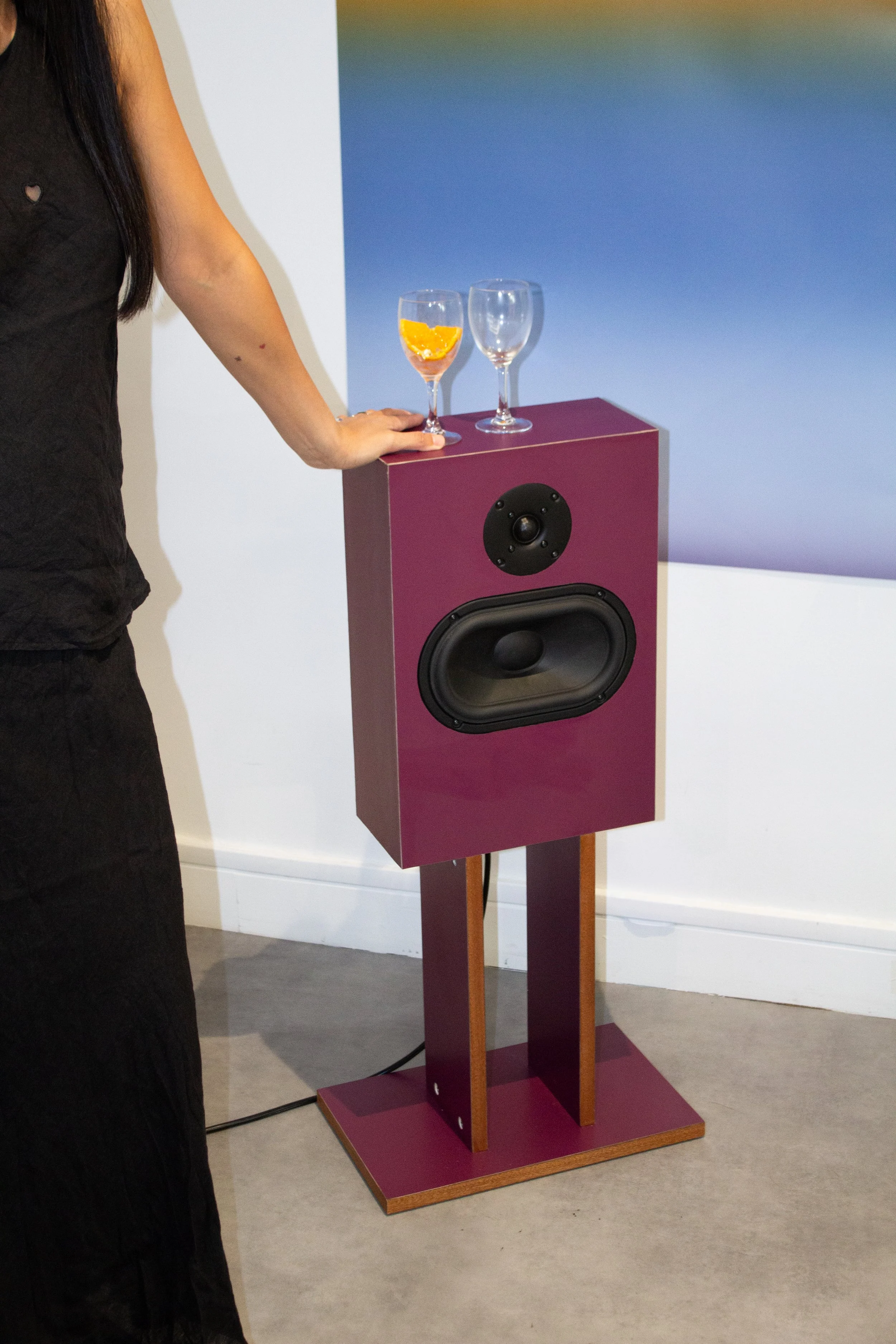 A person standing next to a purple speaker on a stand, with two wine glasses on top, one with orange peel and the other empty, in front of a gradient blue background.