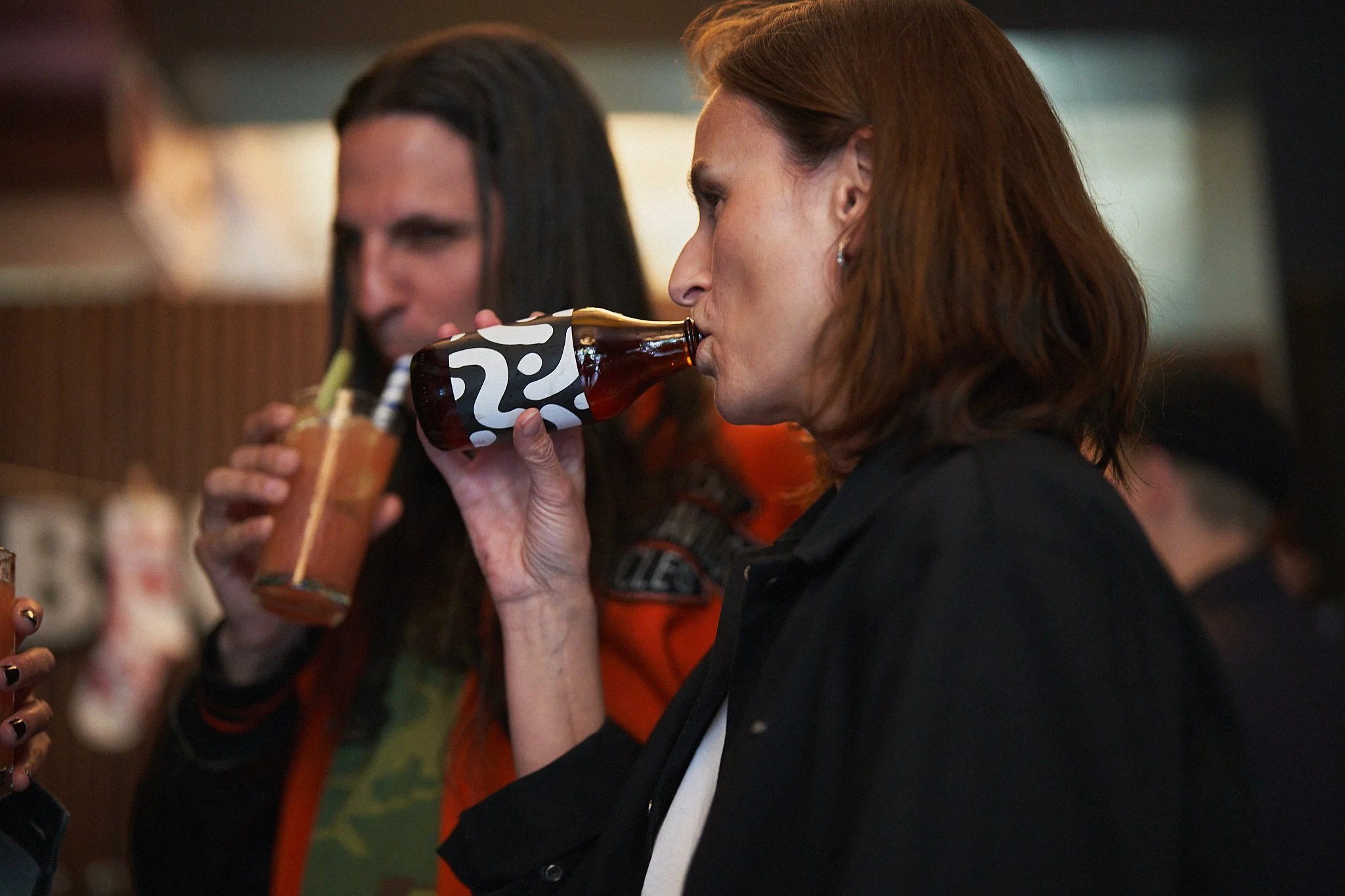 Two women at a bar, one with reddish hair drinking a bottle of scobybaby and the other with long dark hair holding a cocktail.