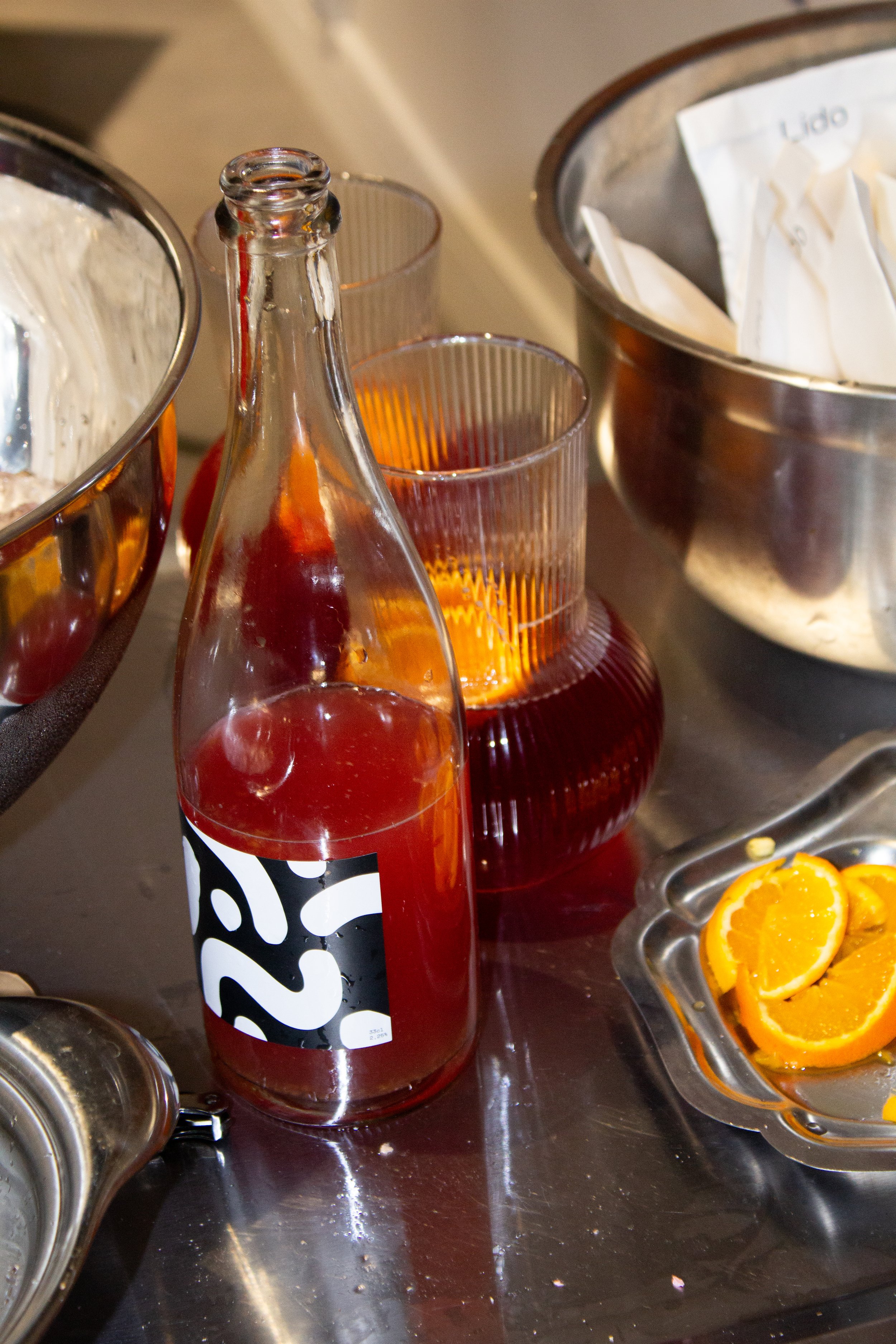 A clear glass bottle with a red scobybaby hibiscus orange (limited edition for Lido), surrounded by orange slices in a metal dish, and empty glasses on a kitchen countertop.