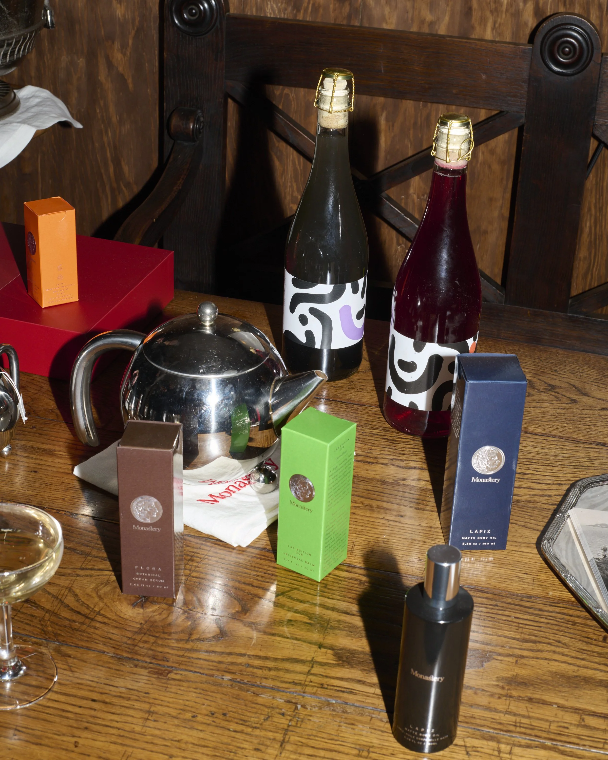 A cluttered wooden table with various beauty and skincare products, including two boxes labeled 'Monastery' in brown and green, a teapot, two bottles of scobybaby, a glass container, and some other small items, with a wooden chair in the background.