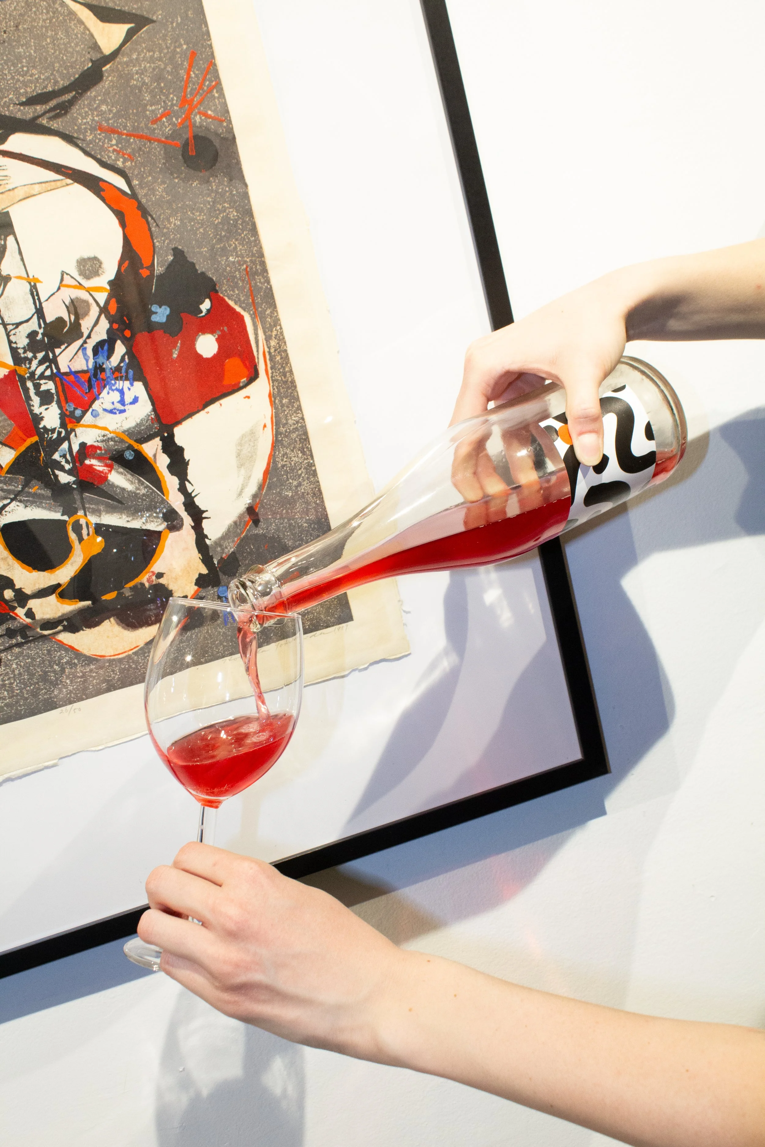 A person is pouring scobybaby kombucha from a bottle with a black and white abstract label into a wine glass. In the background, there is a framed art piece featuring an abstract portrait with bold colors and lines.