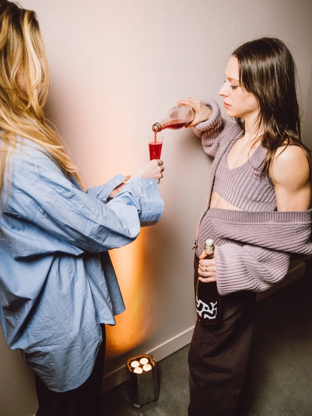 Two women at a party, one pouring a red-colored scobybaby drink into a glass, the other holding a beer bottle, with a small light on the floor.