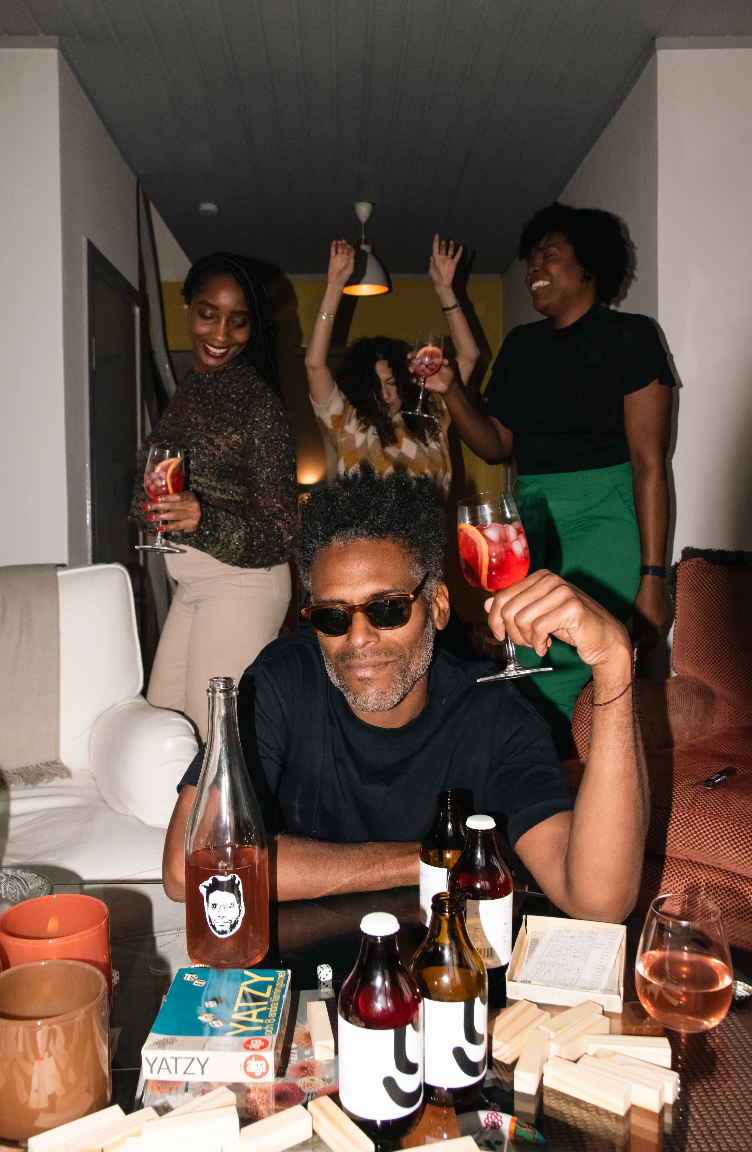 A group of people celebrating indoors, holding drinks, with one person wearing sunglasses and others smiling or dancing, around a table with drinks and games.