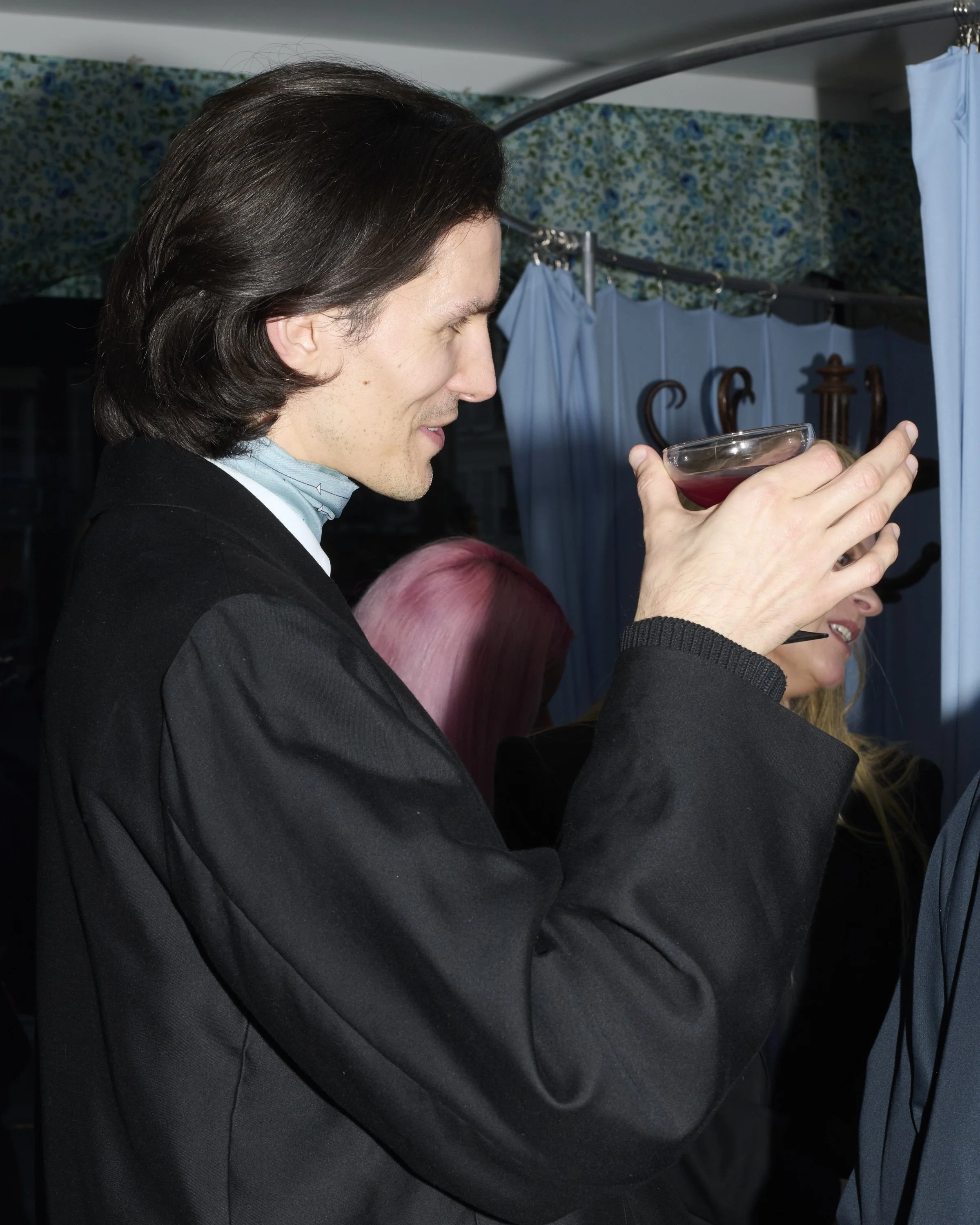 A man with long dark hair, wearing a black suit, holding a drink, standing in a room with blue and green floral wallpaper and blue curtains.