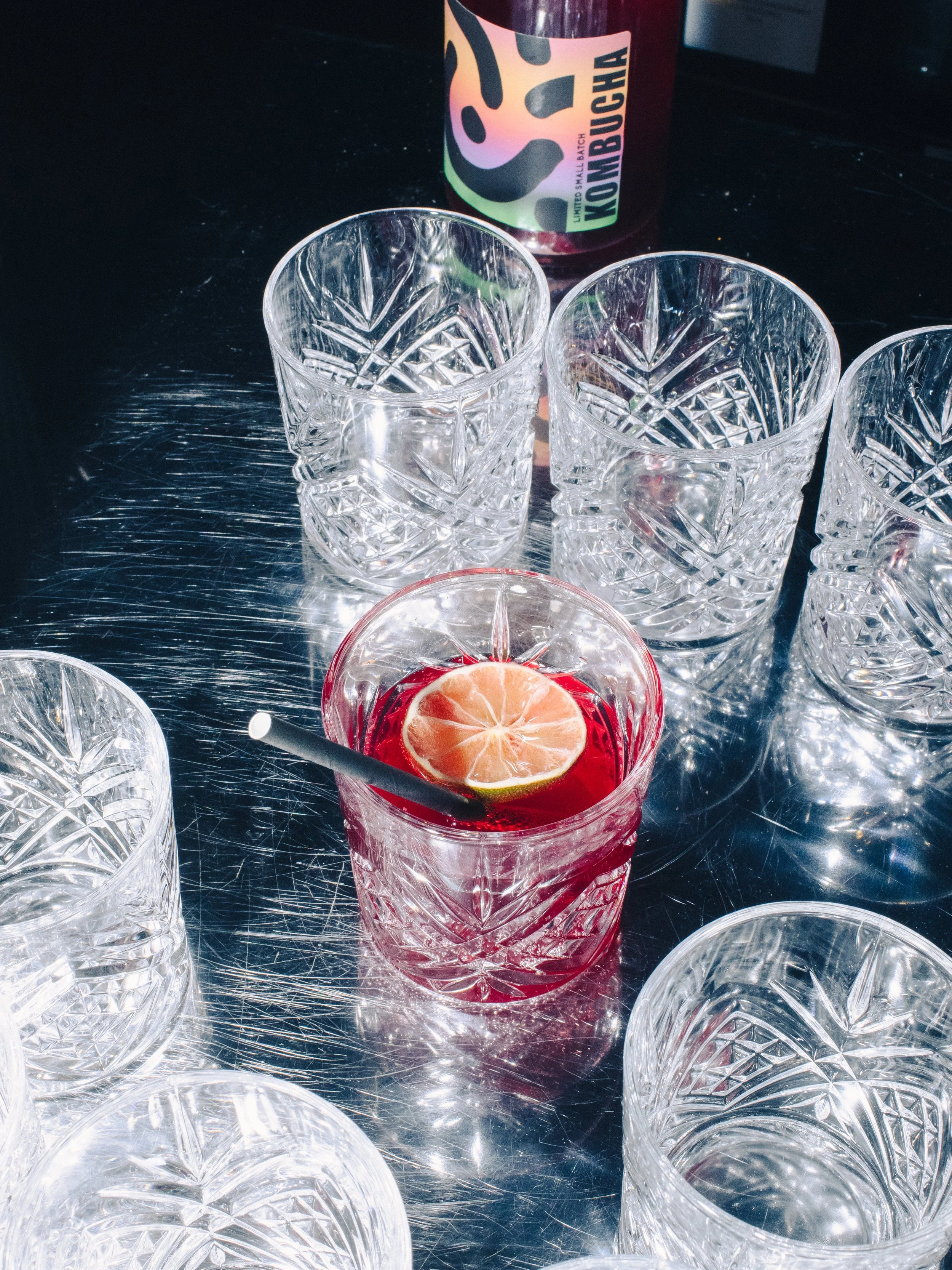 Clear cut-glass tumblers with intricate geometric patterns on a metallic surface. One tumbler contains a slice of scobybaby hibiscus rose kombucha, with a straw, and there is a bottle with colorful abstract design behind the glasses.