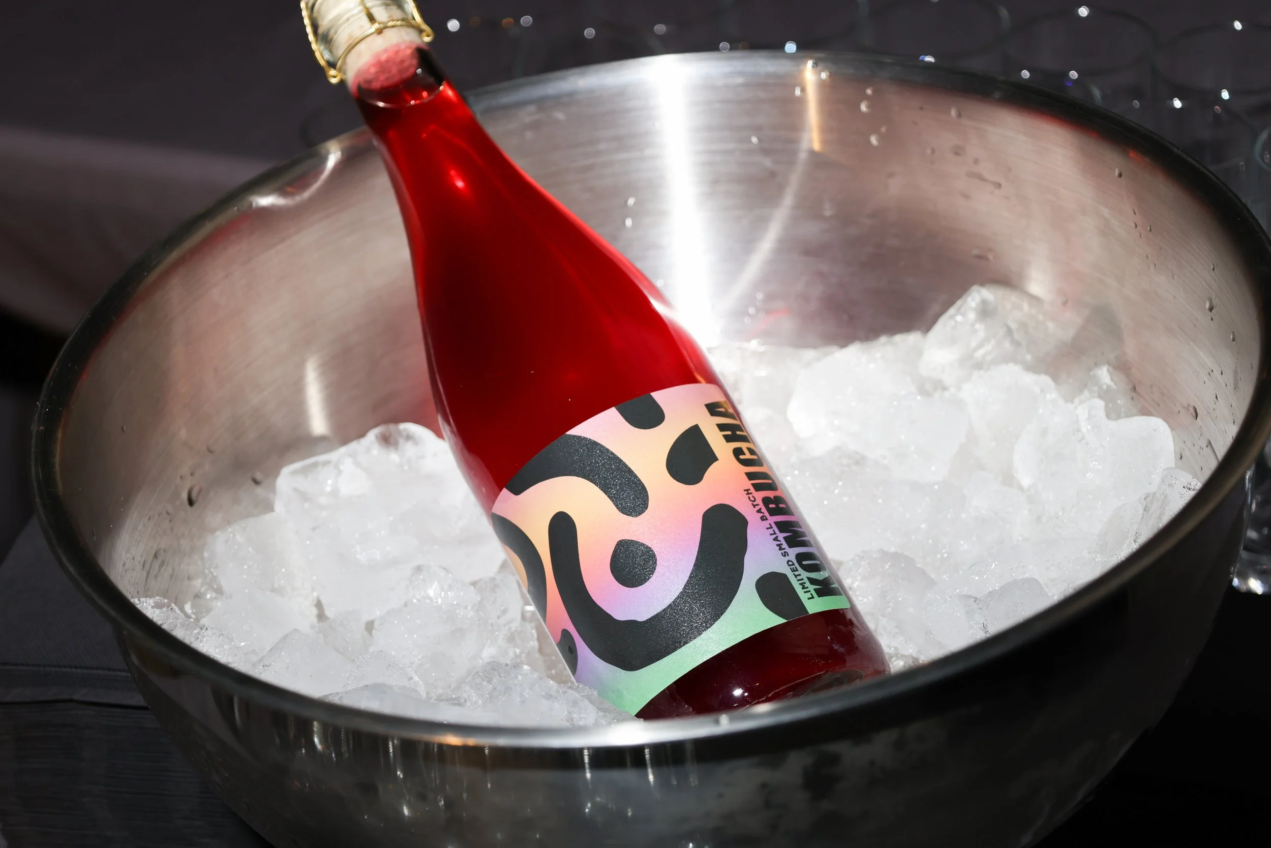 A bottle of scobybaby bissap kombucha with a colorful, abstract label resting on ice inside a metal container, with small ice pieces scattered inside.
