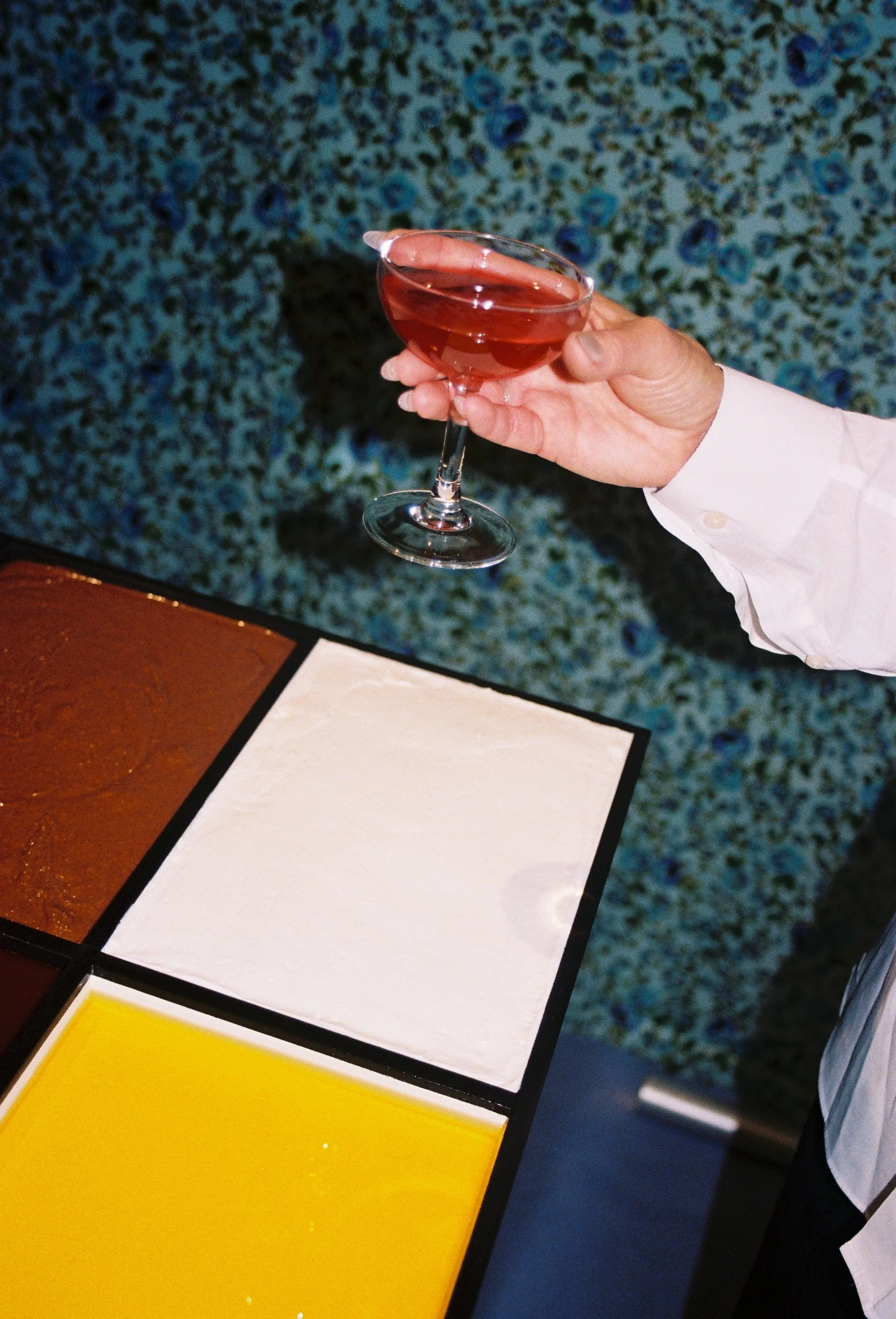 Hand holding a glass of pink liquid, standing near color sample tiles on a table.