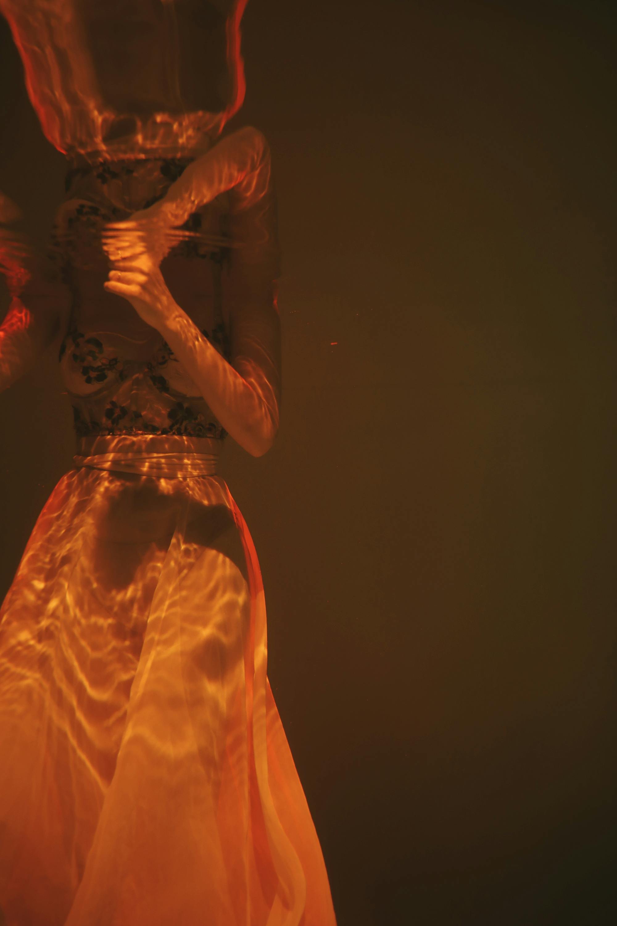 A woman standing in water at night, illuminated by orange and red reflections from a fire or light source, with a dark background.