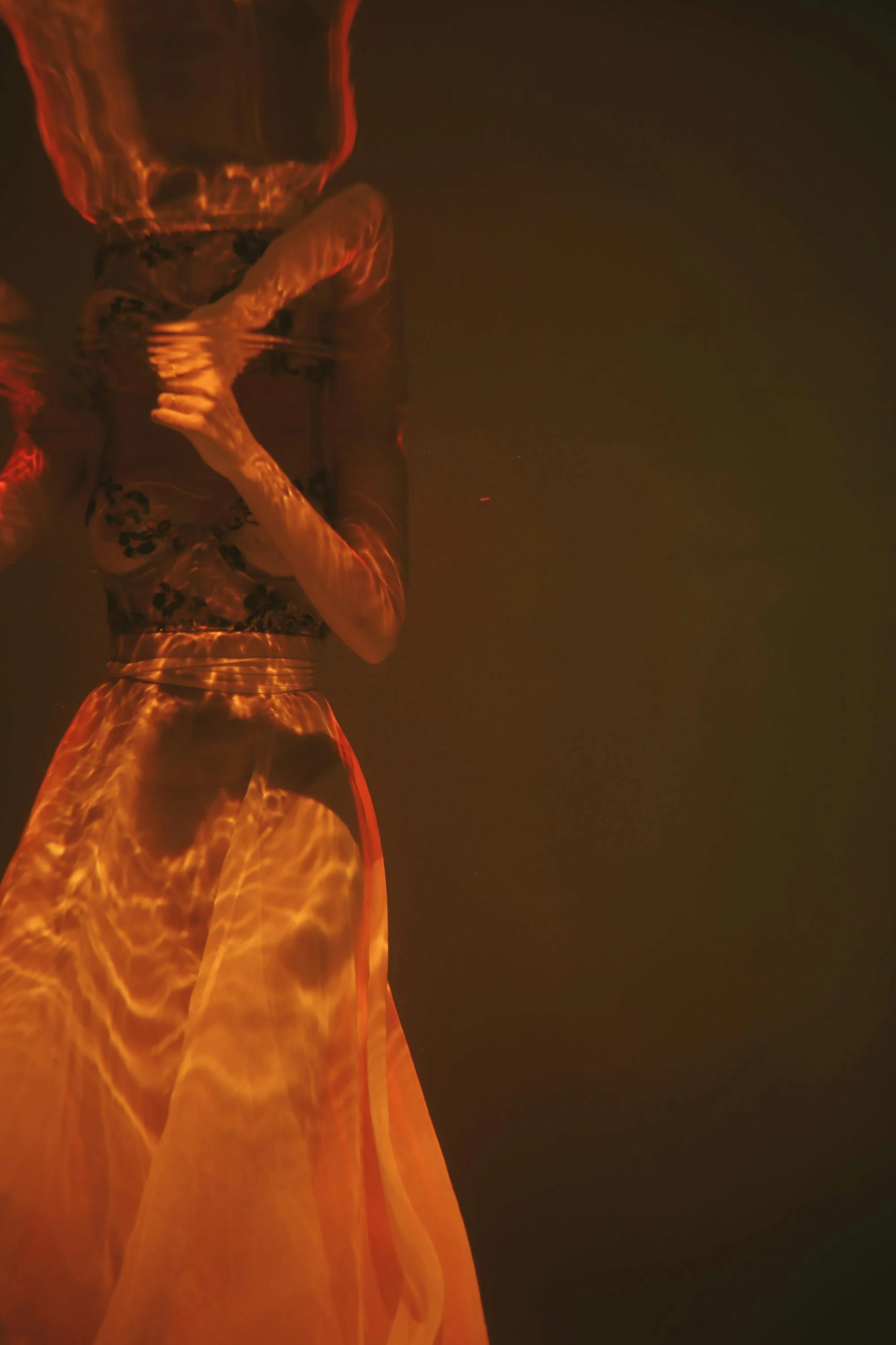 A person wearing a floral patterned dress standing in water that reflects their fiery orange and red dress, with a dark background.