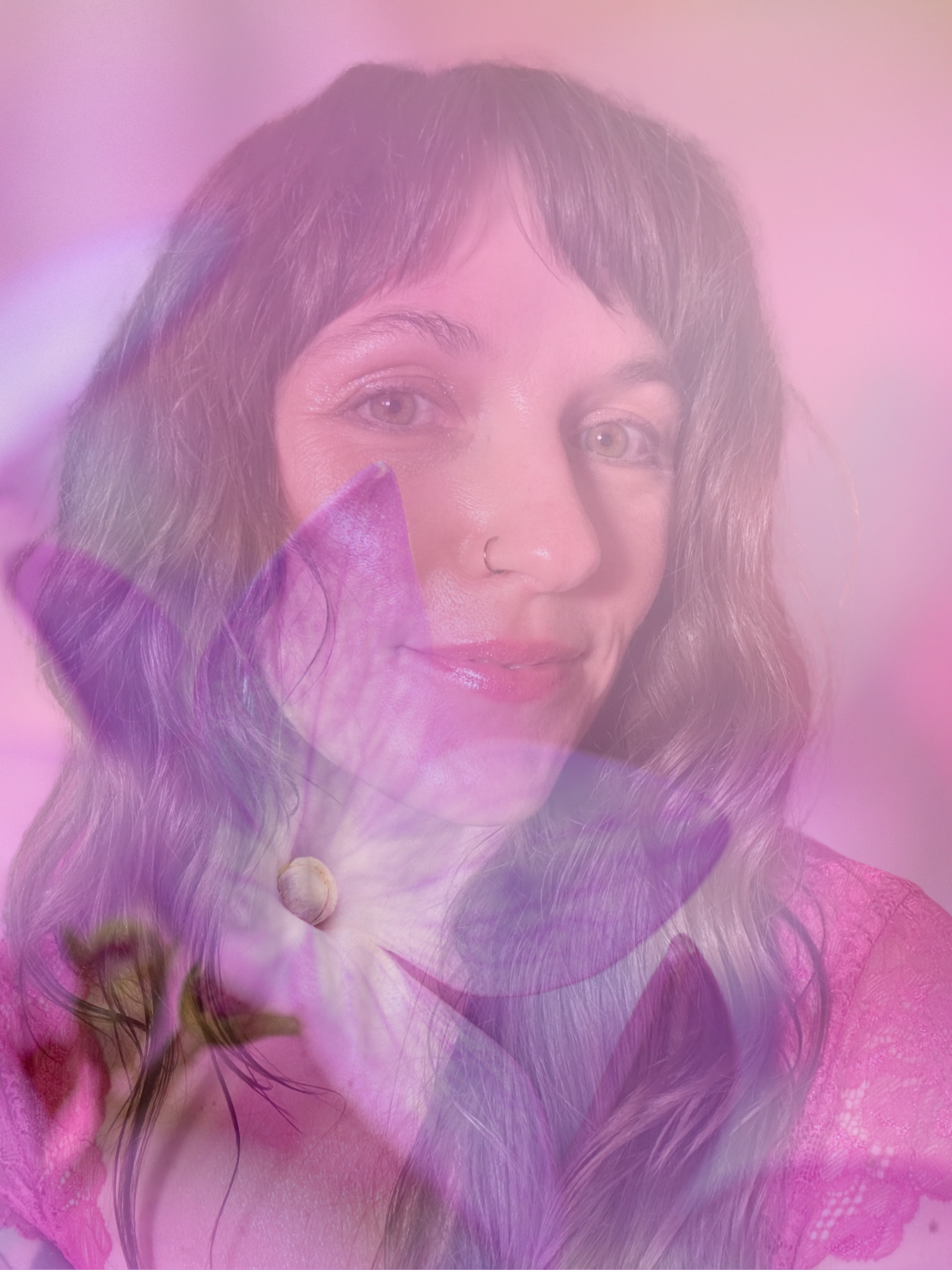 Close-up of a woman with wavy hair and a nose ring, wearing pink lace clothing, with pink and purple floral overlay.