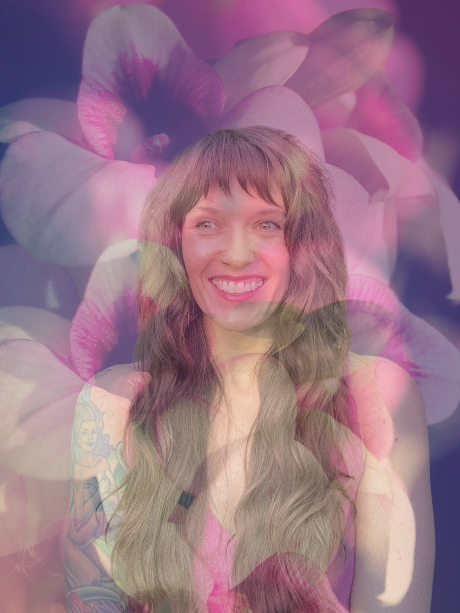 A smiling woman with long brown hair and visible tattoos on her arm, wearing a pink top, with a background of pink and purple flower petals.