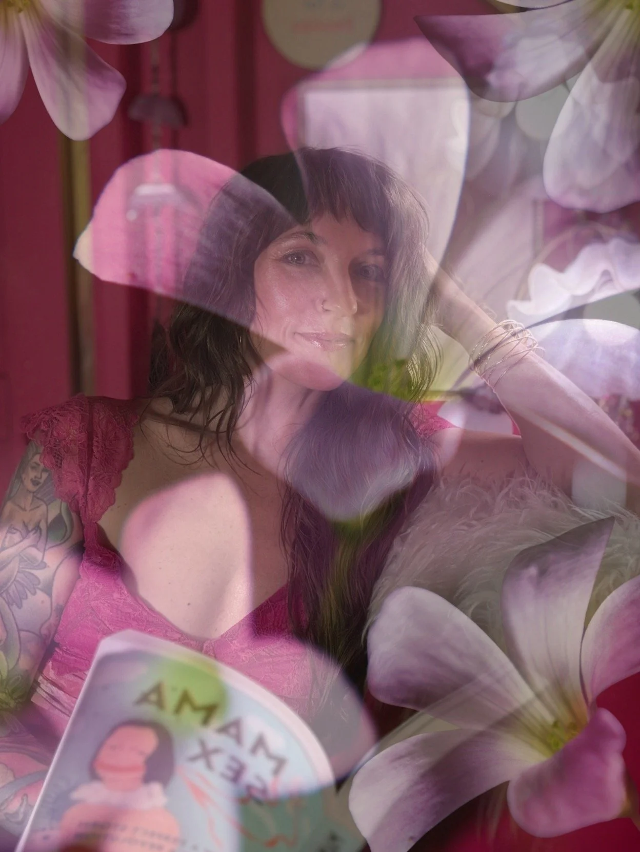 A woman with dark hair and tattoos on her arm, wearing a pink lace top, is surrounded by flower petals or leaves. She is smiling and looking at the camera while holding a card.