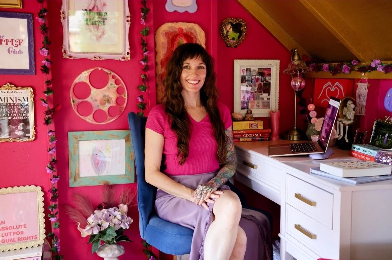 A woman with long brown hair, wearing a pink top and purple skirt, sitting in a blue chair in a colorful, decorated room with pink walls and various framed pictures, books, and decorative items on the desk and wall.