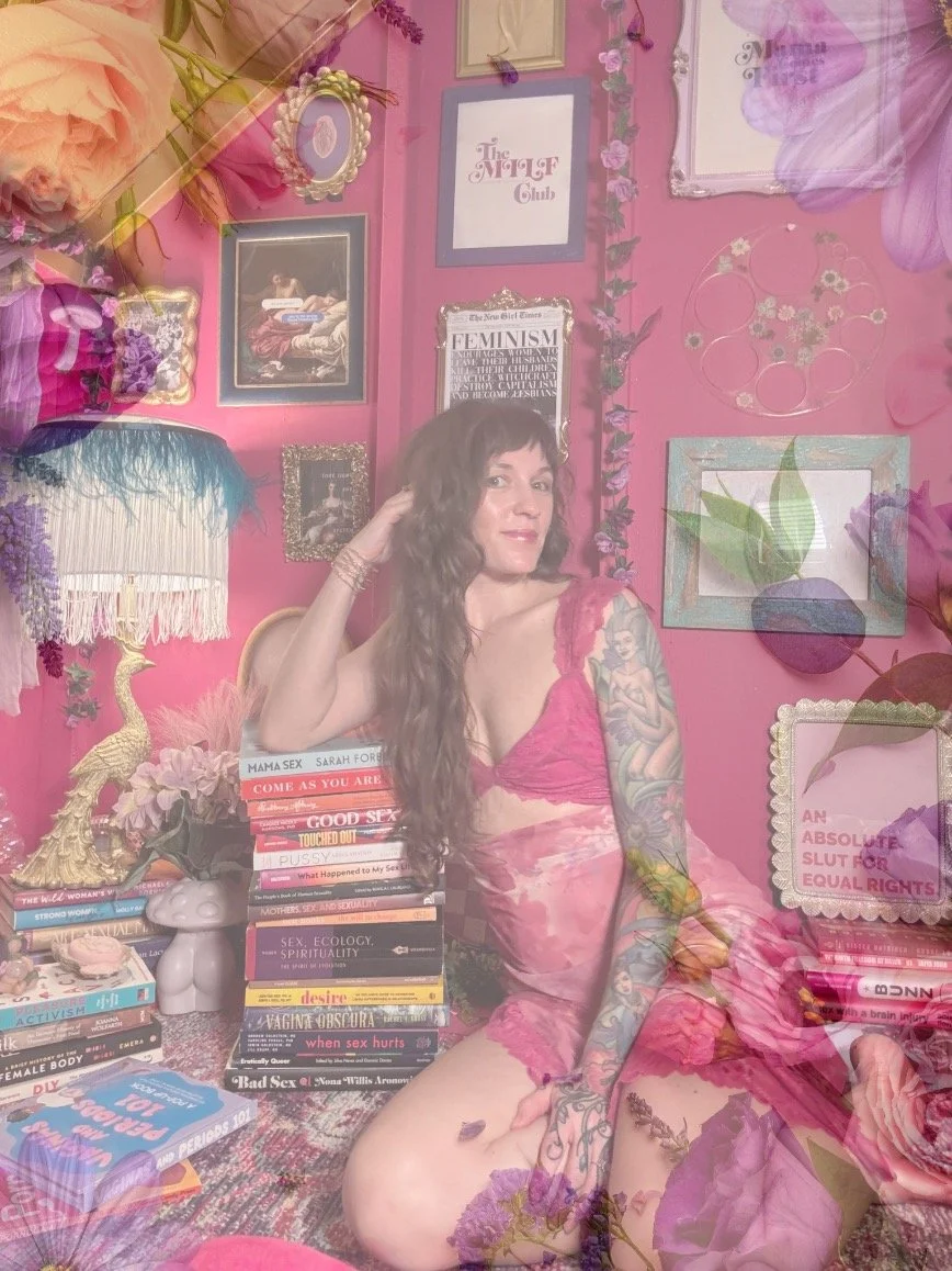 A woman with long curly hair and tattoos, wearing pink lingerie, sitting among a stack of books about sex and feminism in a pink decorated room with framed artwork, flowers, and a stuffed animal.