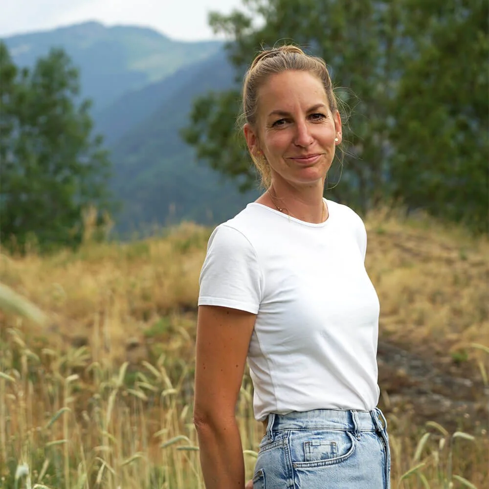 Fabienne Escher, Porträt in der Natur, vor einem Kornfeld in den Bergen, Grengiols, weißes T-Shirt, Jeans. Sie lächelt freundlich in die Kamera.