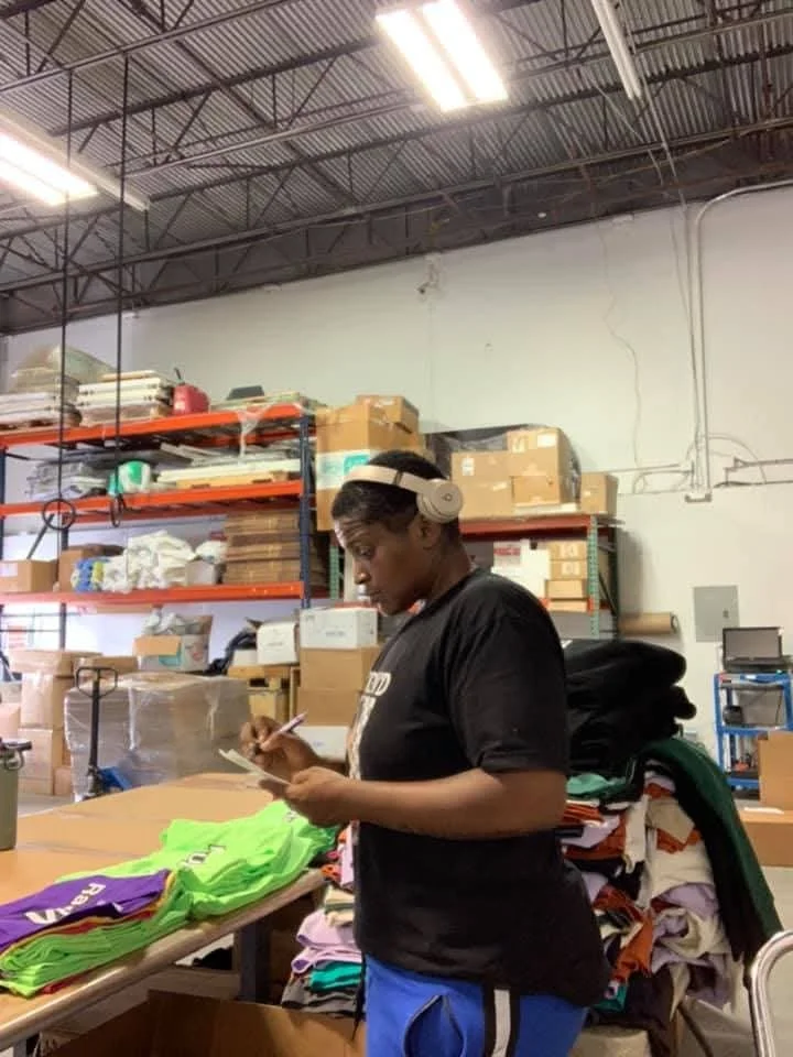 A young woman with short hair wearing headphones, a black T-shirt, and blue shorts, standing at a table in a warehouse, writing on a notepad while surrounded by stacks of colorful T-shirts and shelves filled with boxes and supplies.