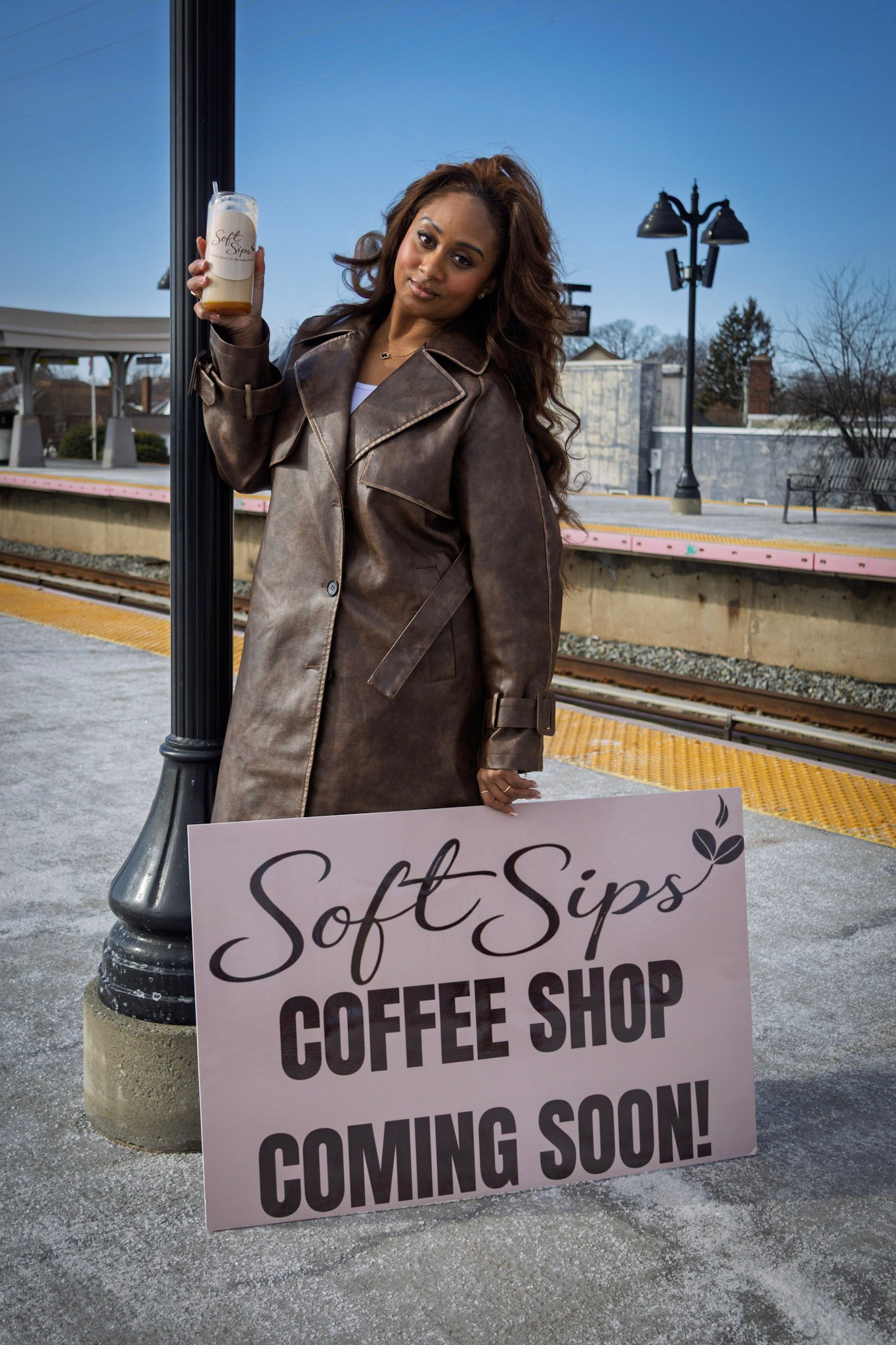 A woman standing outdoors at a train station holding a beverage with a sign that reads "Soft Sips Coffee Shop Coming Soon!"