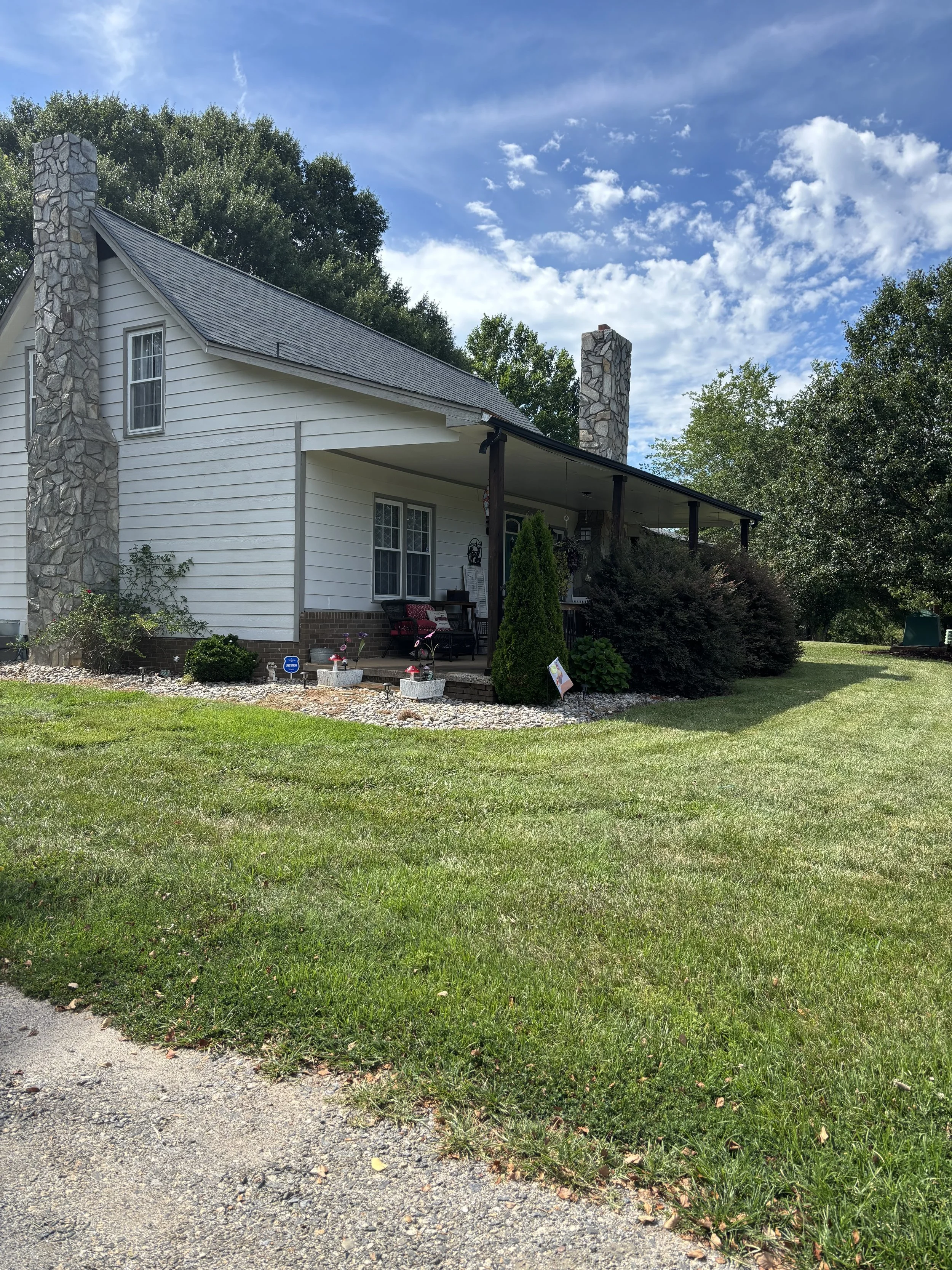 Front yard of a house with a green lawn, small plants, and a gravel pathway under a partly cloudy blue sky.