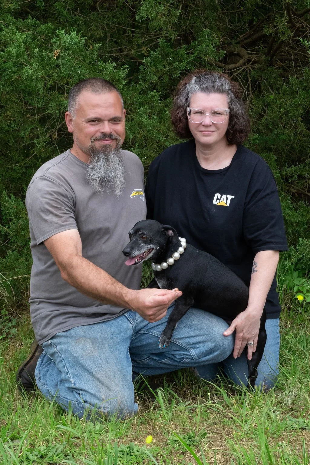 A man and woman kneeling outdoors with a black dog wearing a pearl necklace. Both are smiling, greenery in the background.