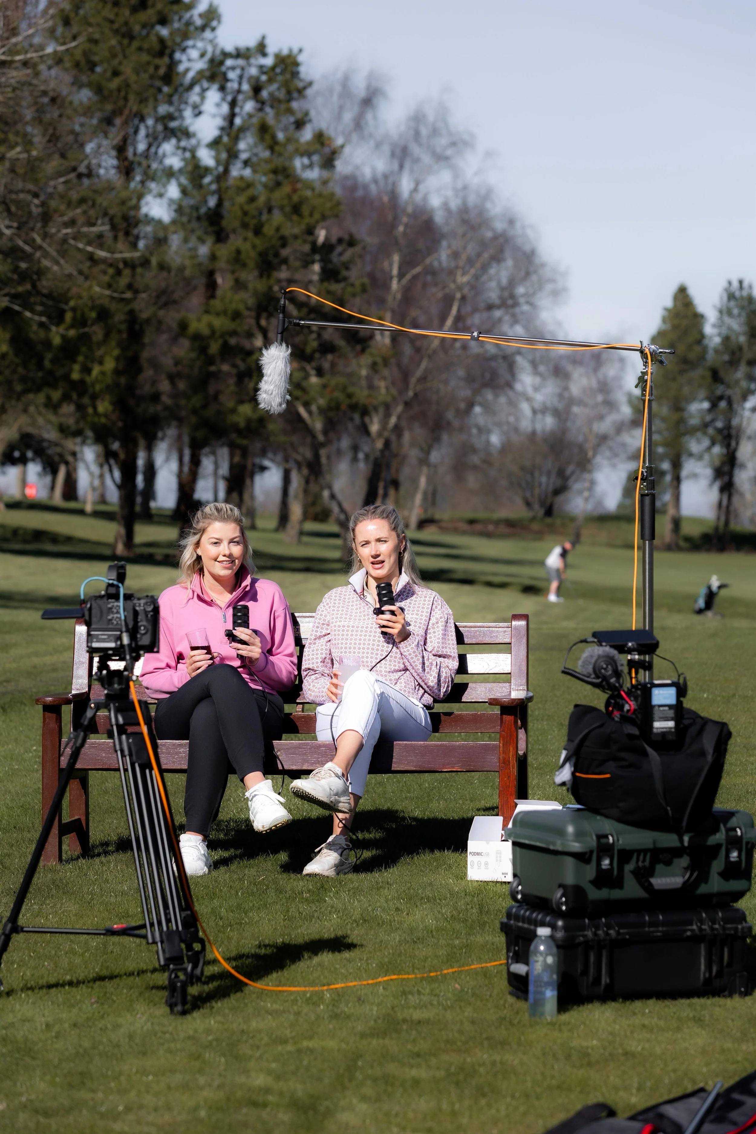 Two women sitting on a park bench surrounded by filming equipment, including cameras, microphones, and lighting, with trees and a person in the background.