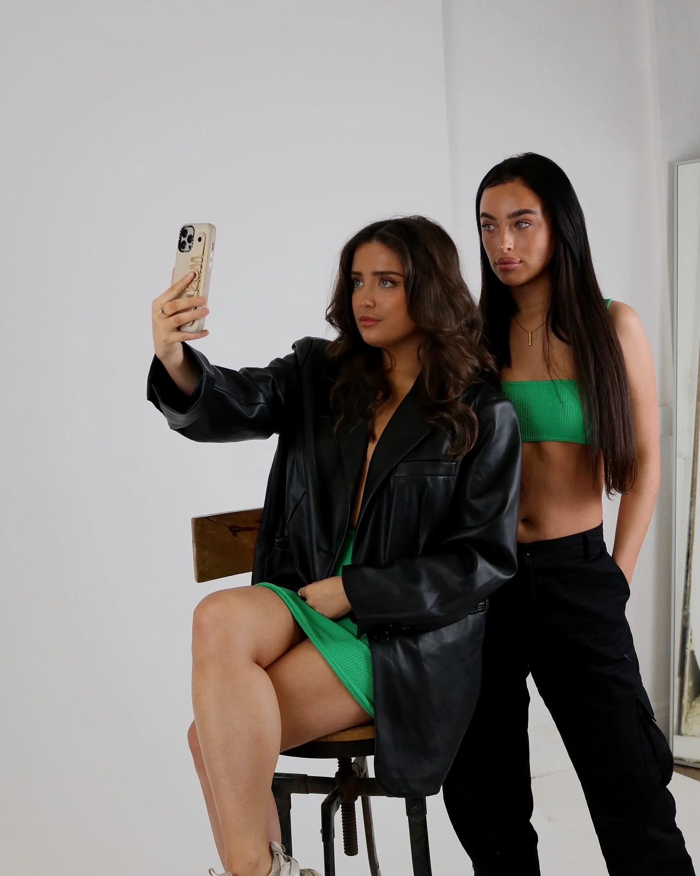 Two women posing for a photo, one sitting on a stool and the other standing behind her, both looking at the camera. The woman sitting is wearing a green dress and a black leather jacket, and the woman standing is wearing a green top and black pants.