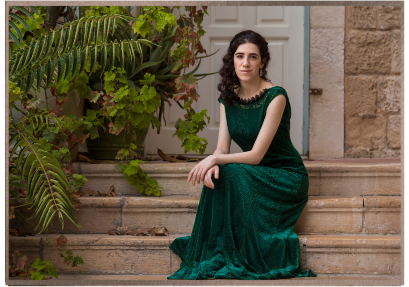 Portrait of Adi Denner seated on historic stone steps surrounded by greenery, wearing a green gown.