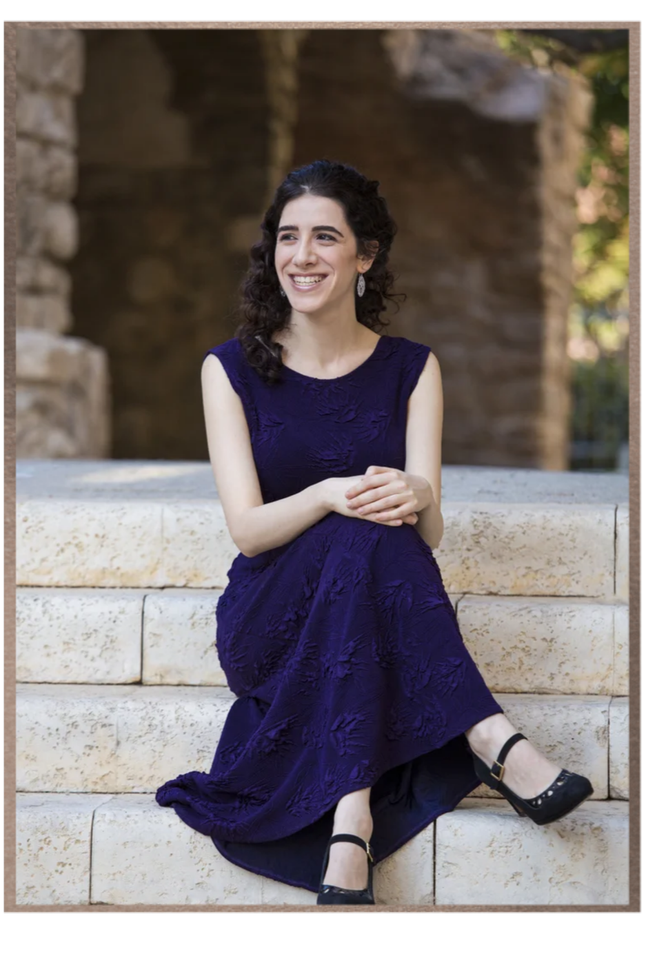 Portrait of  Adi Denner seated on stone steps in a deep purple dress, smiling.