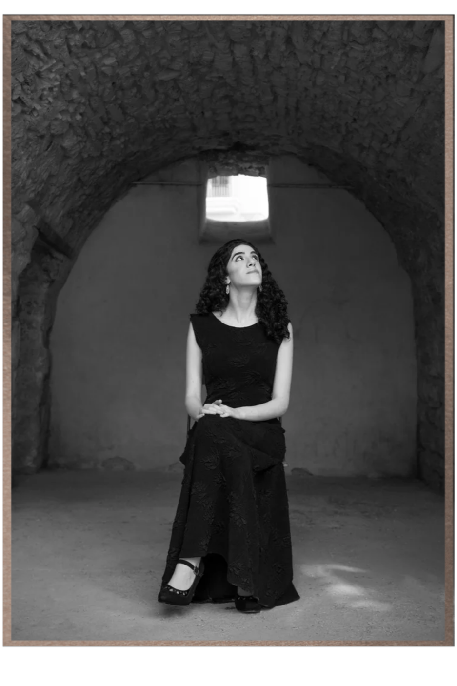 Black-and-white portrait of Adi Denner standing beneath a stone archway and looking toward the light.