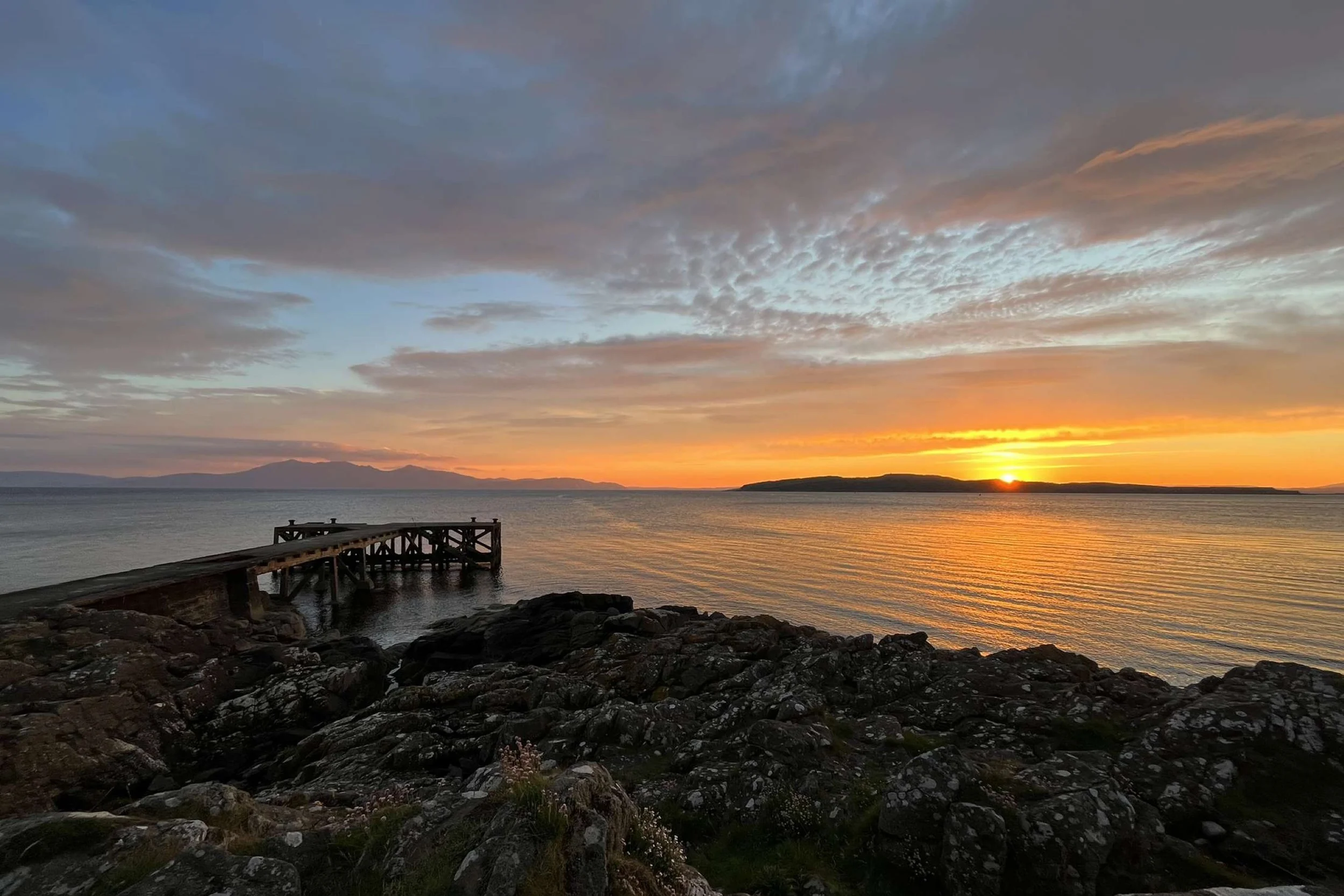 Sunset over a body of water with a rocky shoreline and a wooden pier in the foreground, cloudy sky with hues of orange and blue.