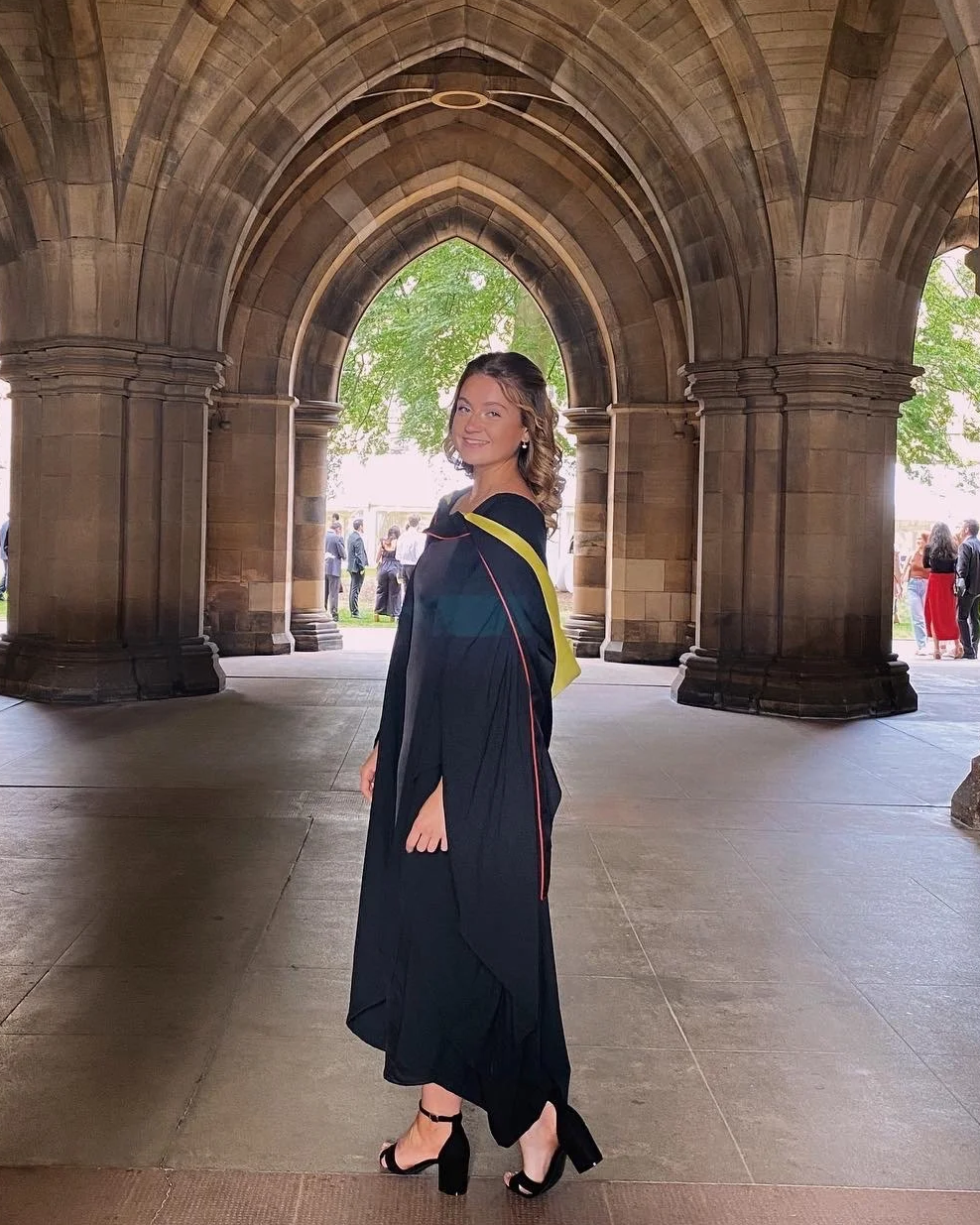 A young woman in a graduation gown standing under a stone archway, smiling at the camera, with students and trees in the background.