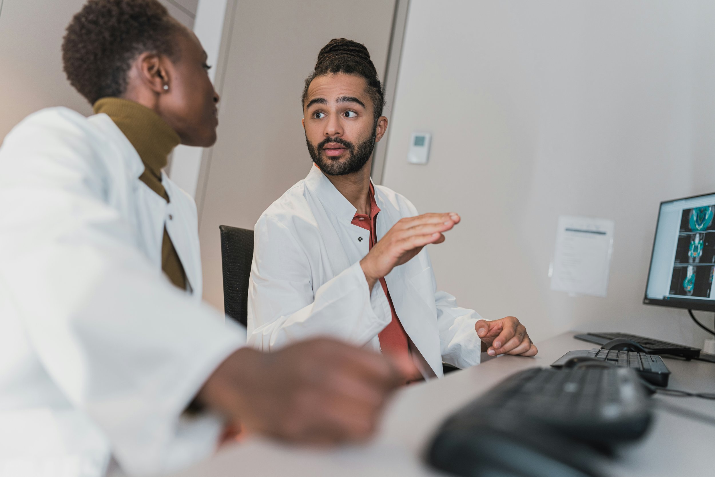Deux médecins discutent devant un ordinateur dans un hôpital ou un laboratoire, avec des images médicales visibles à l'écran.