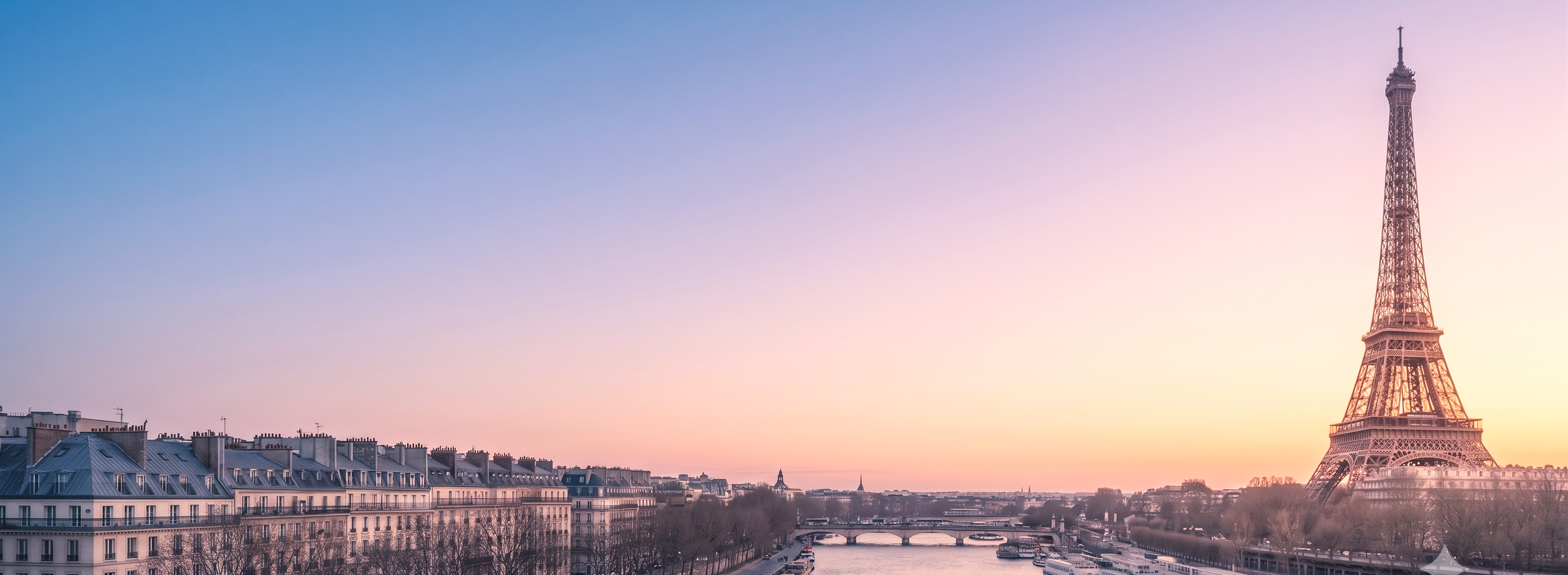 Coucher de soleil sur la ville de Paris avec la Tour Eiffel visible à droite, bâtiments résidentiels et la Seine avec un pont au premier plan.