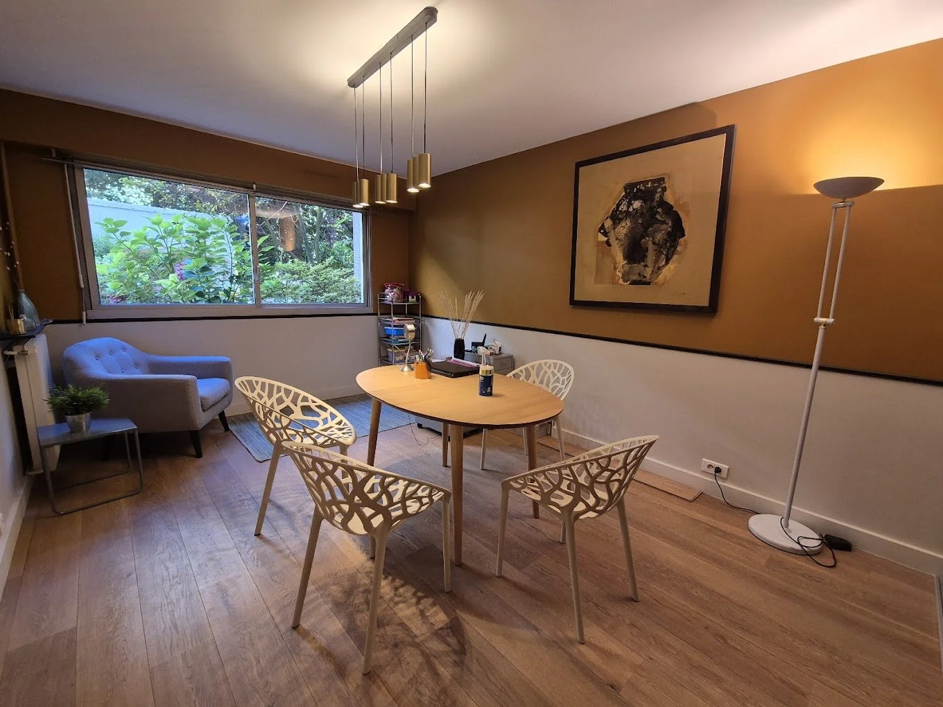 Salle à manger avec un bureau rond en bois, quatre chaises en plastique blanc avec motif, un fauteuil beige, une étagère en métal, une lampe sur pied, une fresque artistique, une grande fenêtre donnant sur un jardin vert.