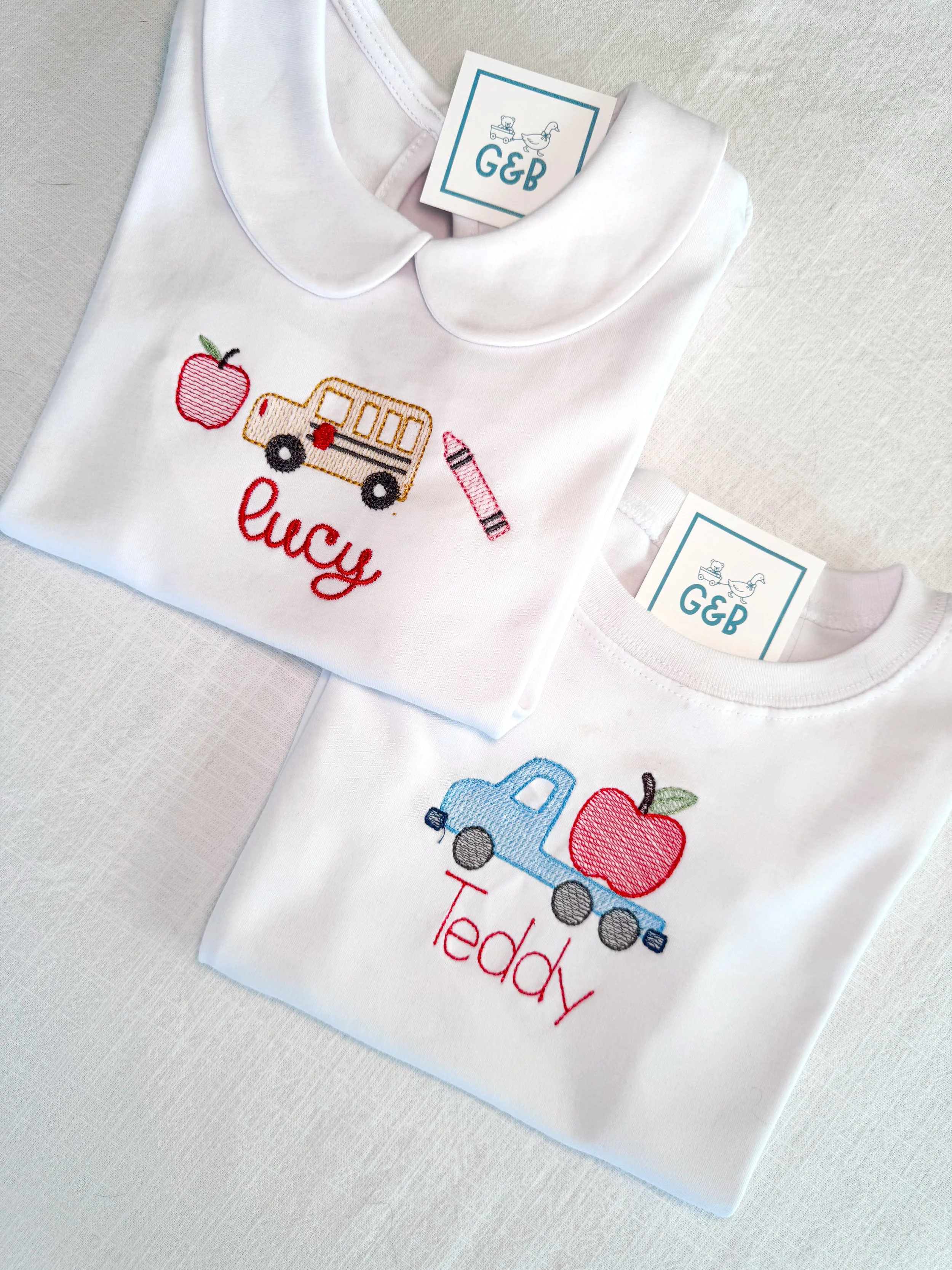 Two white toddler T-shirts with embroidered designs, one with a school bus, apple, and crayon and the other with a toy dump truck and apple. Both shirts have colorful embroidered text.