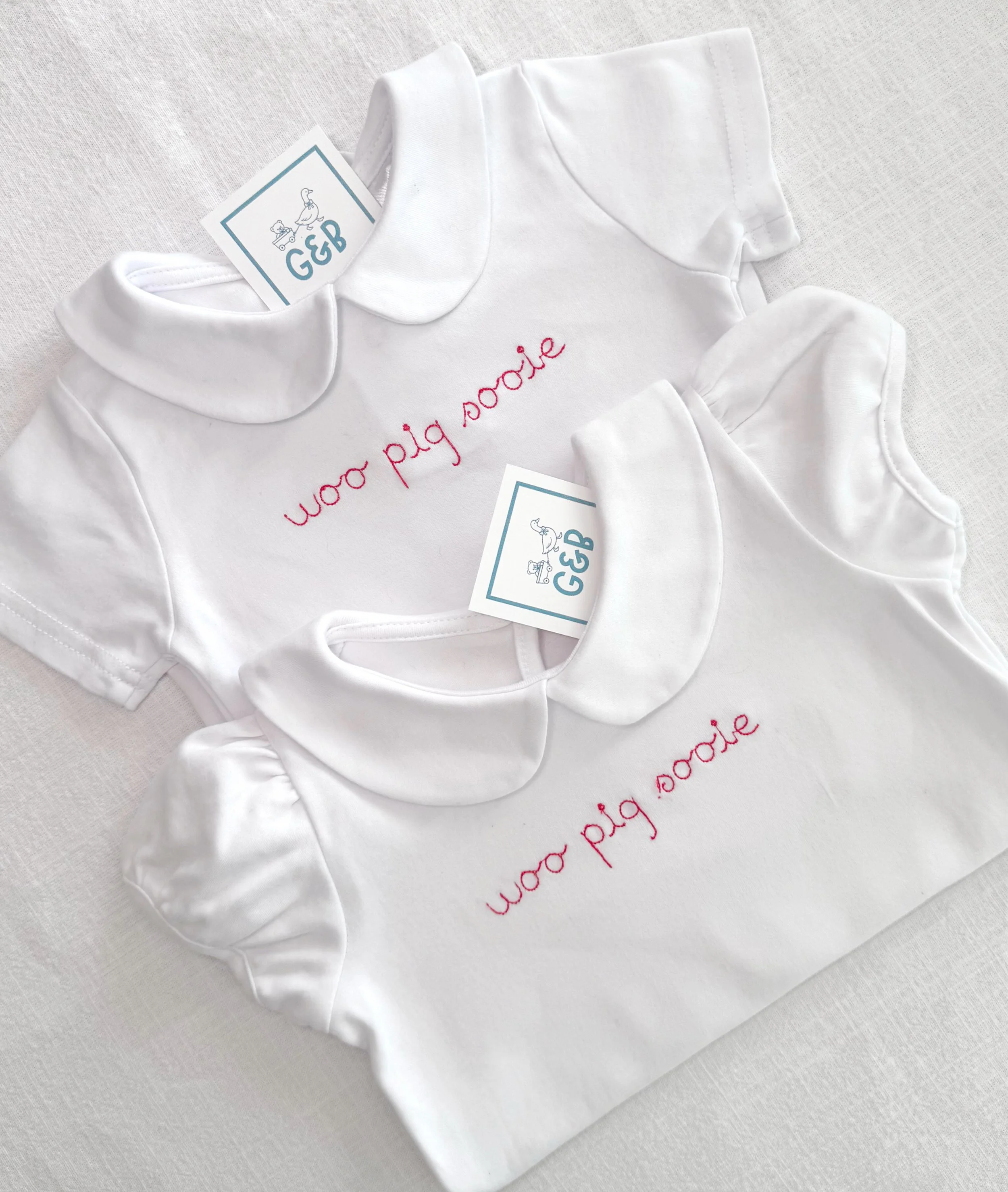 Baby white onesie with pink embroidered text and a label from G&B featuring a duck and a bear illustration.