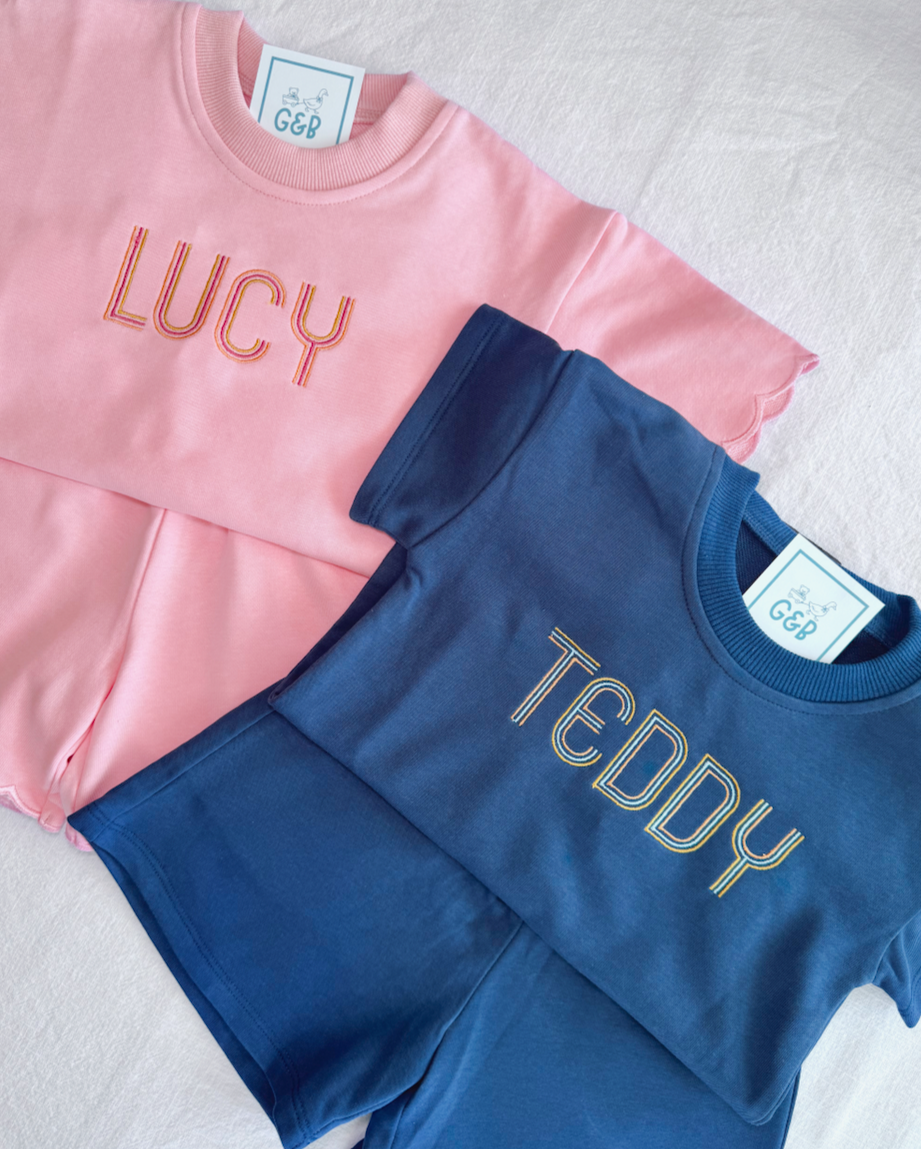 Pink sweatshirt with 'LUCY' embroidered on it, and blue sweatshirt with 'TEDDY' embroidered on it, both laid flat on a light fabric surface.