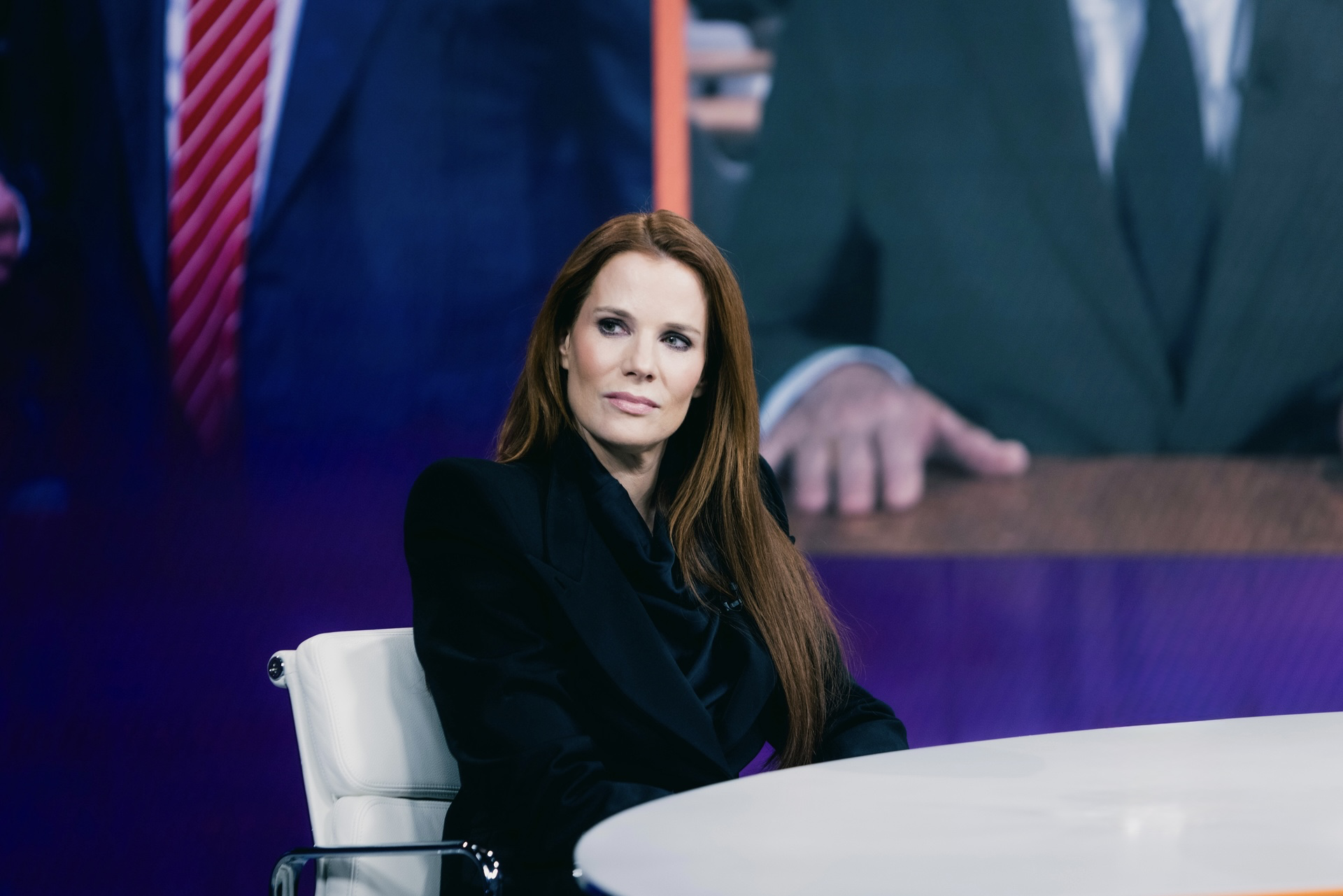 A woman with long red hair, wearing a black blazer, sitting at a conference table with a serious expression. In the background, there is a large screen displaying a man in a suit, partially visible.