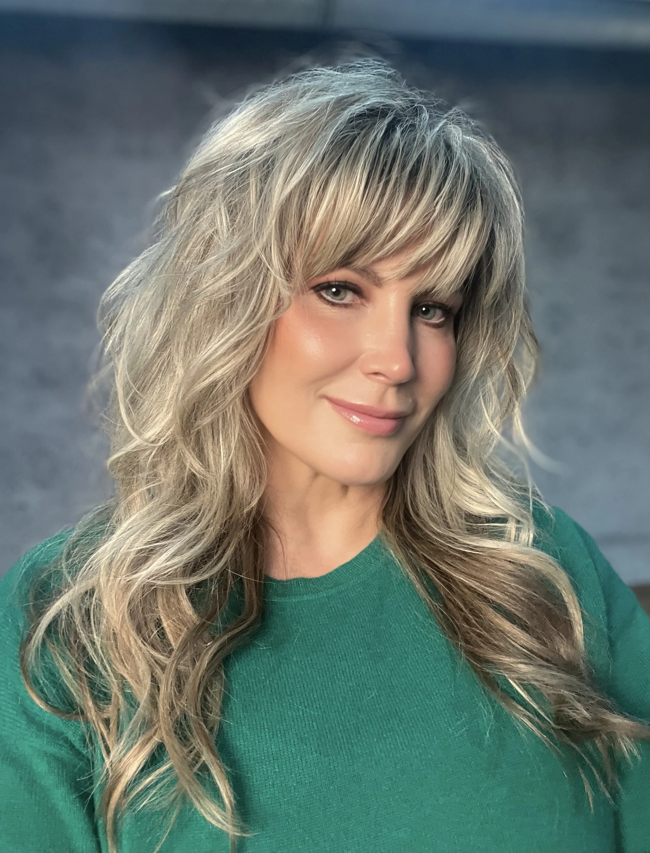 Close-up of a woman with long, wavy blonde hair wearing a green top, with a blurred gray background.