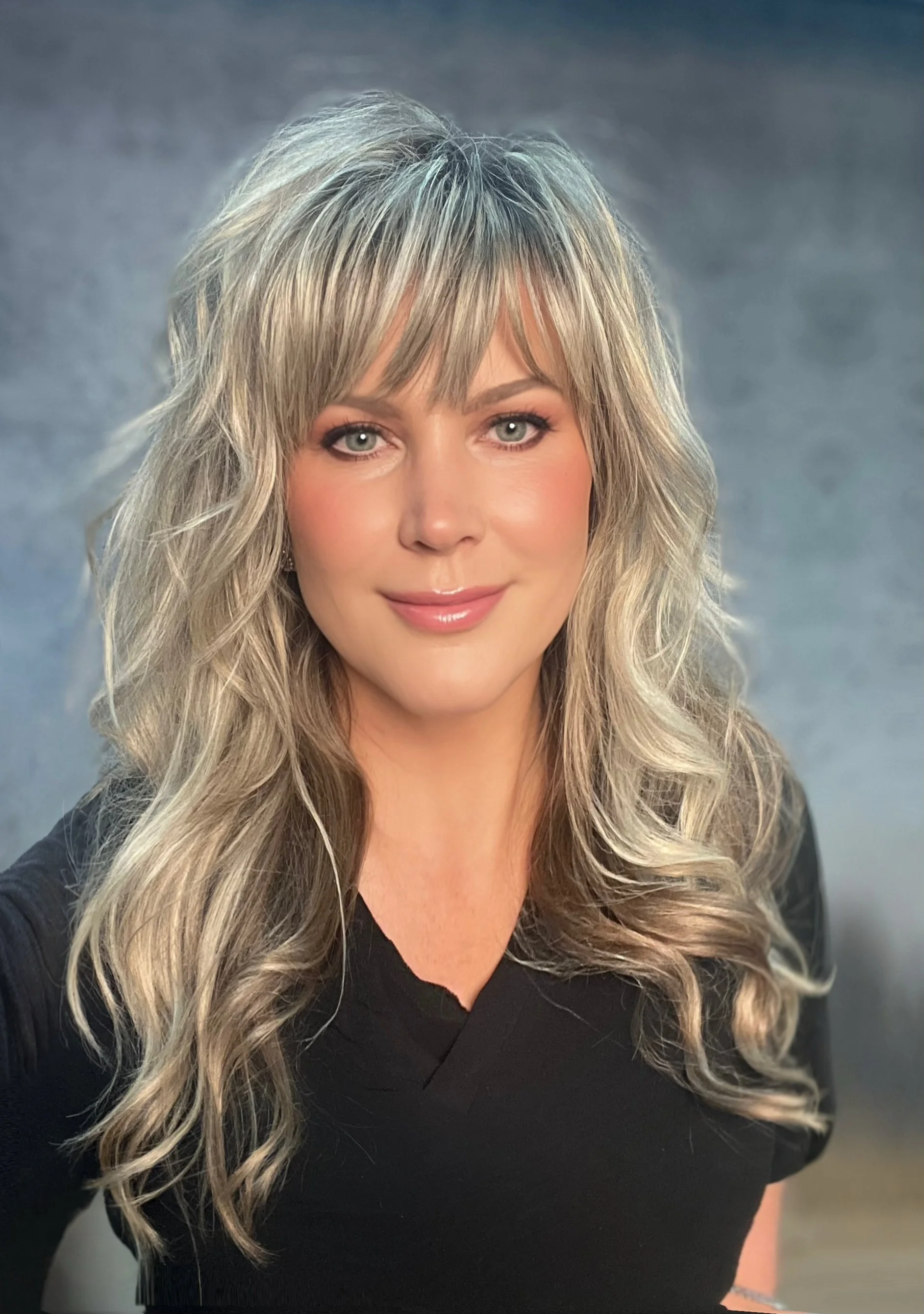 Close-up portrait of a woman with long, wavy blonde hair, light makeup, and a black top, smiling softly against a neutral background.