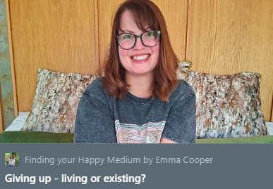 A young woman with shoulder-length red hair, glasses, and a smile, sitting on a bed in a room with wooden paneled walls, wearing a gray shirt with a patterned chest design.