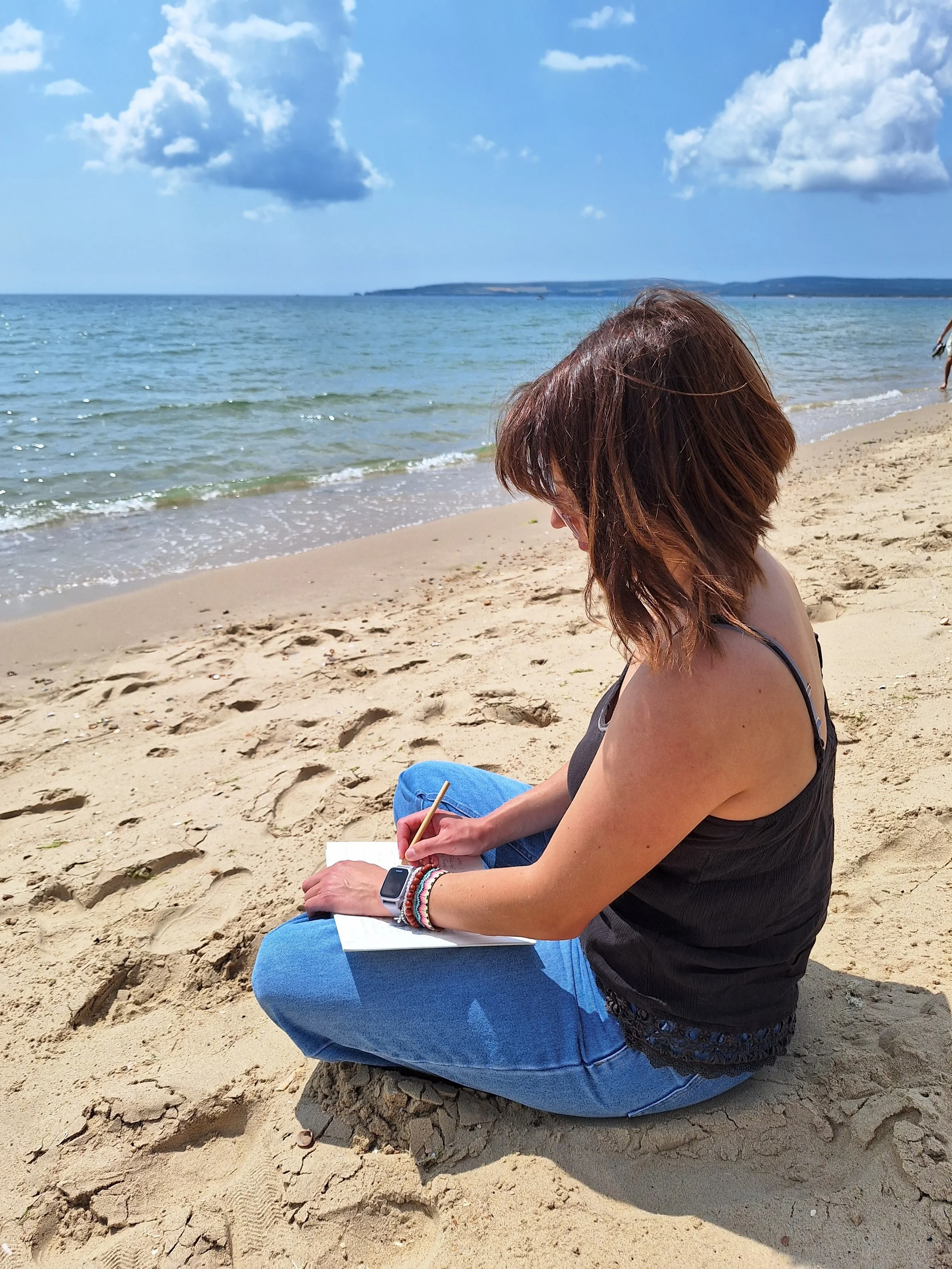 A woman with brown hair, wearing a black tank top and blue jeans, sitting on a rock on a sandy beach, writing in a notebook with a pencil, with the ocean and a cloudy sky in the background.