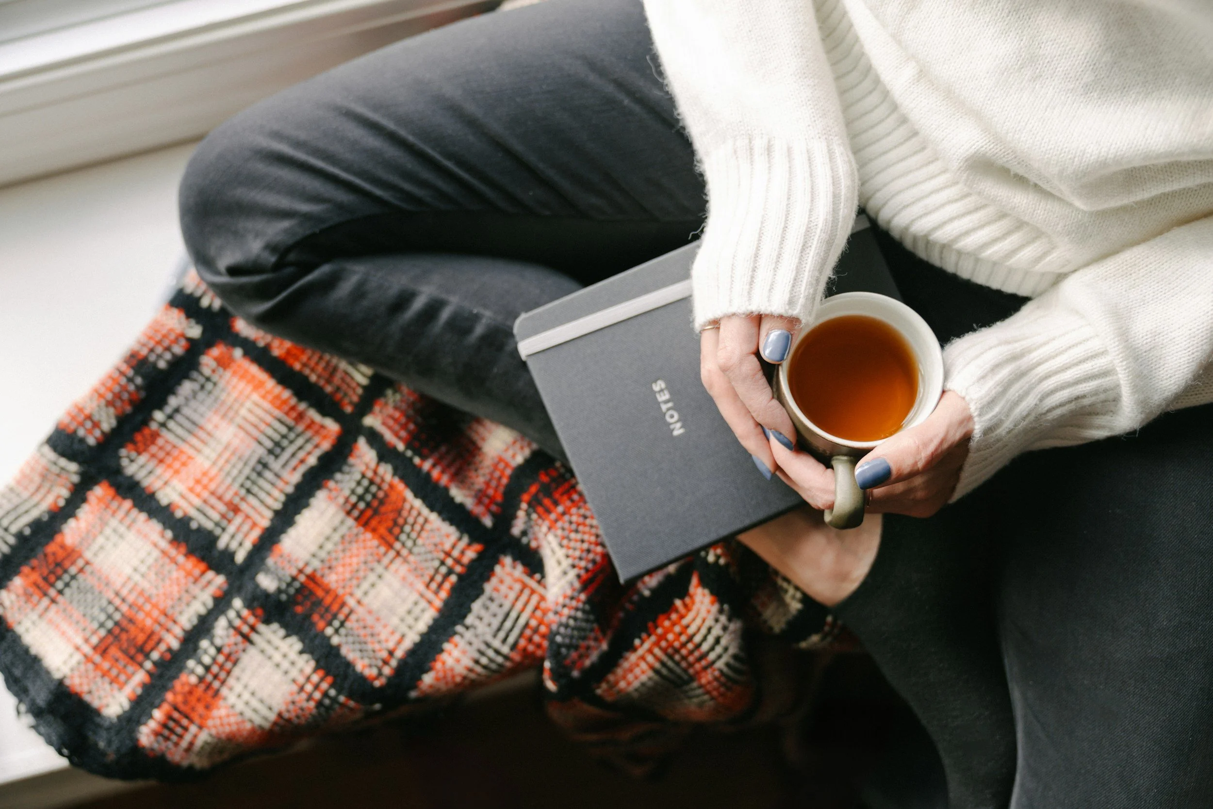Person sitting by a window, holding a cup of tea or coffee, wearing a cream-colored sweater and dark pants, with a notebook or journal on their lap, and a colorful patterned blanket over their legs.